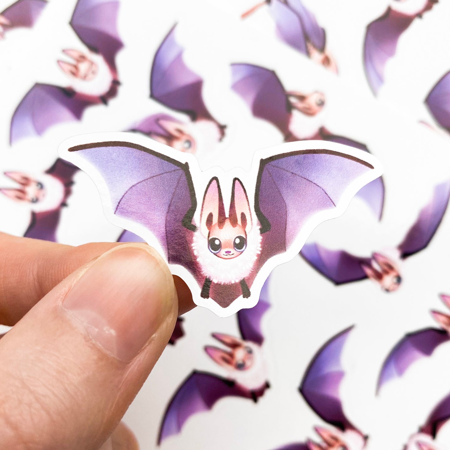 A sticker of a flying bat with its wings stretched out. Each sticker is an individual unique drawing of a bat in a different pose. Most bats are flying, but one is hanging upside down from a banch and sleeping with it's face tucked inside its wings. While another is sitting down to rest.