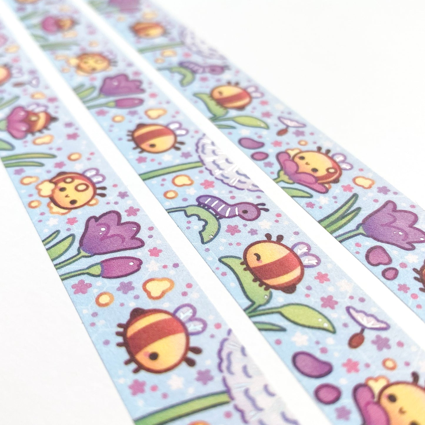 A different angle showing the holographic silver foil reflecting in the light. The foil is used to enhance some features, like making the bee wings more shiny and adding highlights to the plants. The caterpillar has holographic stripes!