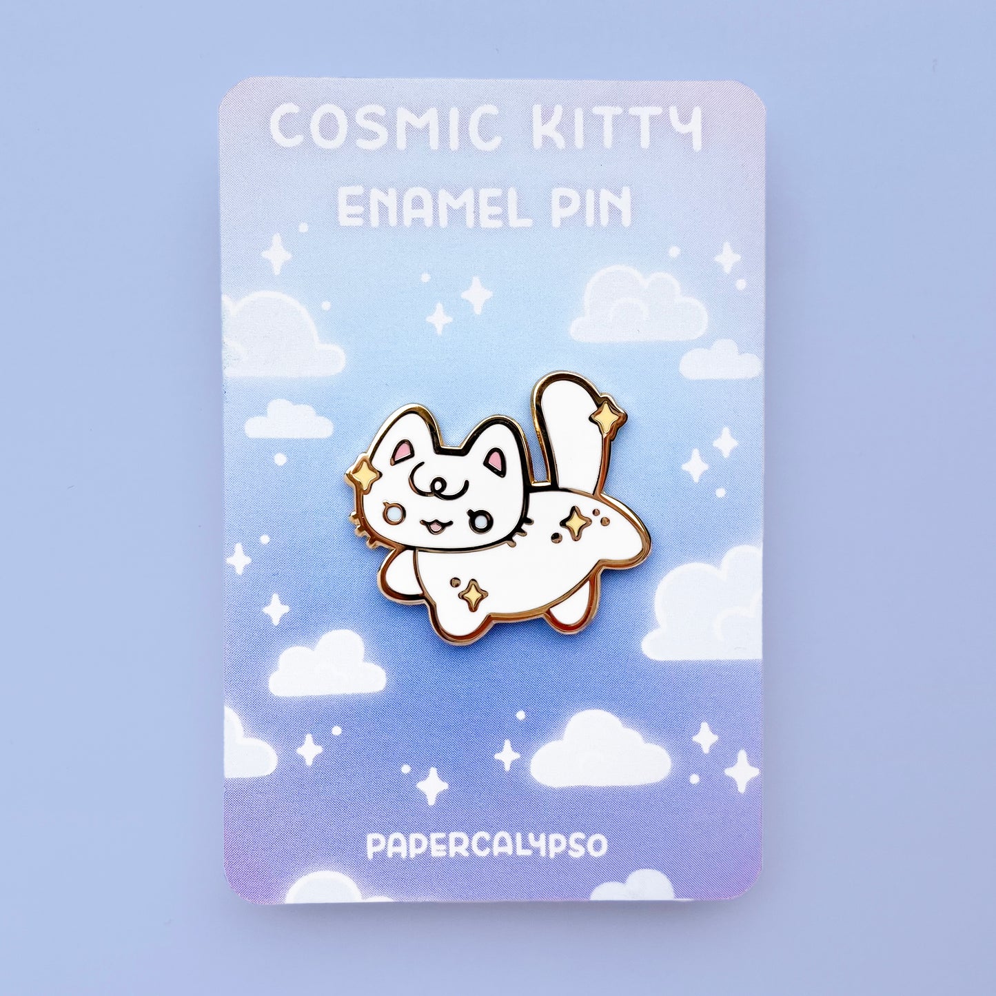 A gold hard enamel pin of a white kitty cat with stars in its fur. The stars are yellow and it has pretty light blue eyes. It's striking a cute pose by lifting one hind leg and one front leg and smiling.