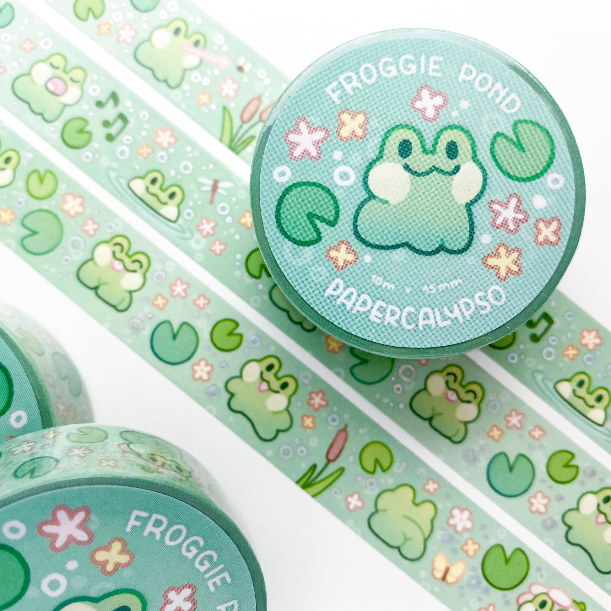 kawaii frog themed washi tape featuring green cartoon froggies on a blue background with lily pads. The frogs are happily jumping, singing, swimming and having a great time together. One is even showing us his little butt!