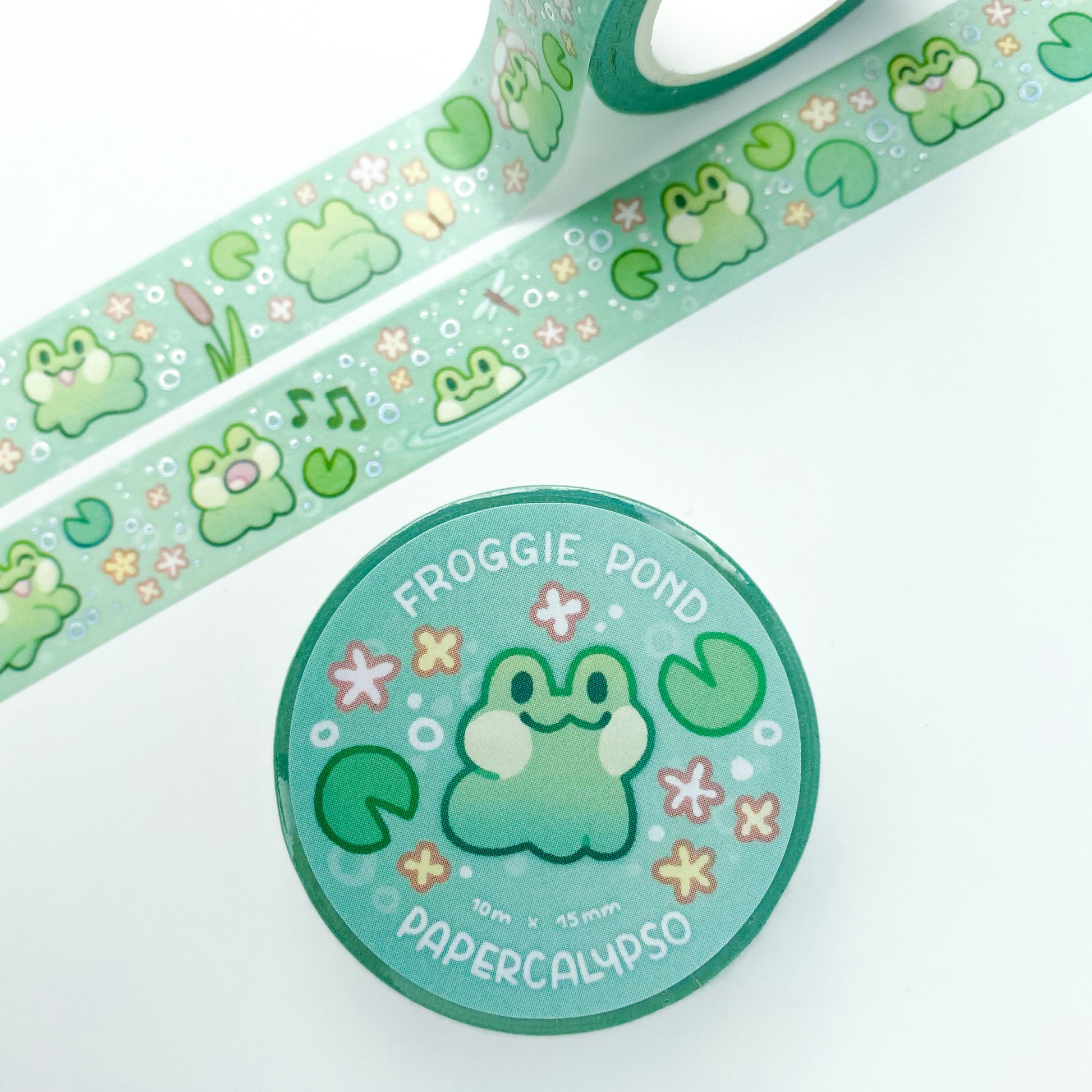 the roll of washi tape is packaged with shrink wrap plastic and a round sticker on one side. On it is an adorable smiling frog surrounded by flowers and lily pads. It reads "Froggie Pond, 10 metres by 15 milimetres, by Papercalypso"