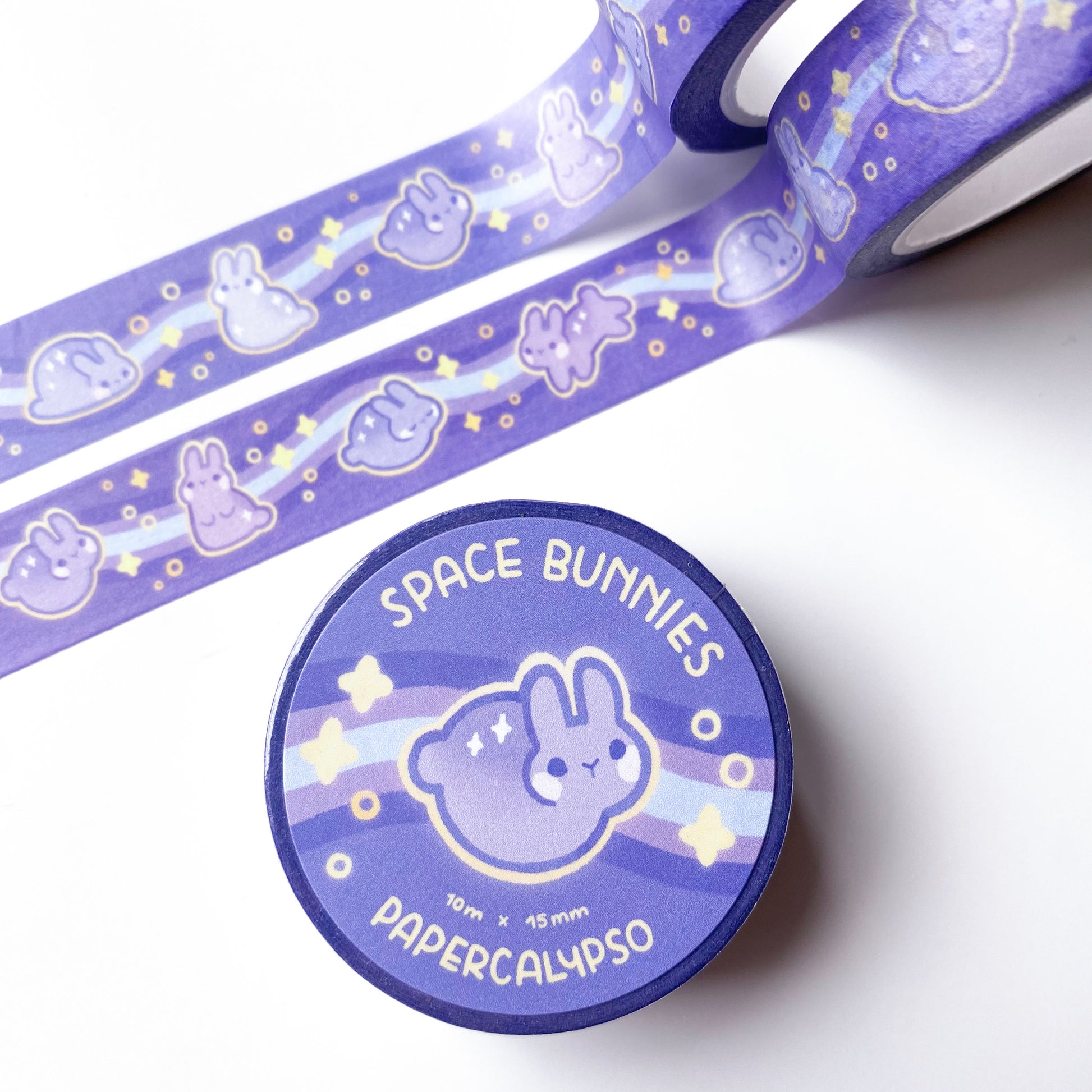 the roll of washi tape is packaged with shrink wrap plastic and a round sticker on one side. On it is a round space rabbit without legs, surrounded by yellow stars. It reads "Space Bunnies, 10 metres by 15 milimetres, by Papercalypso"