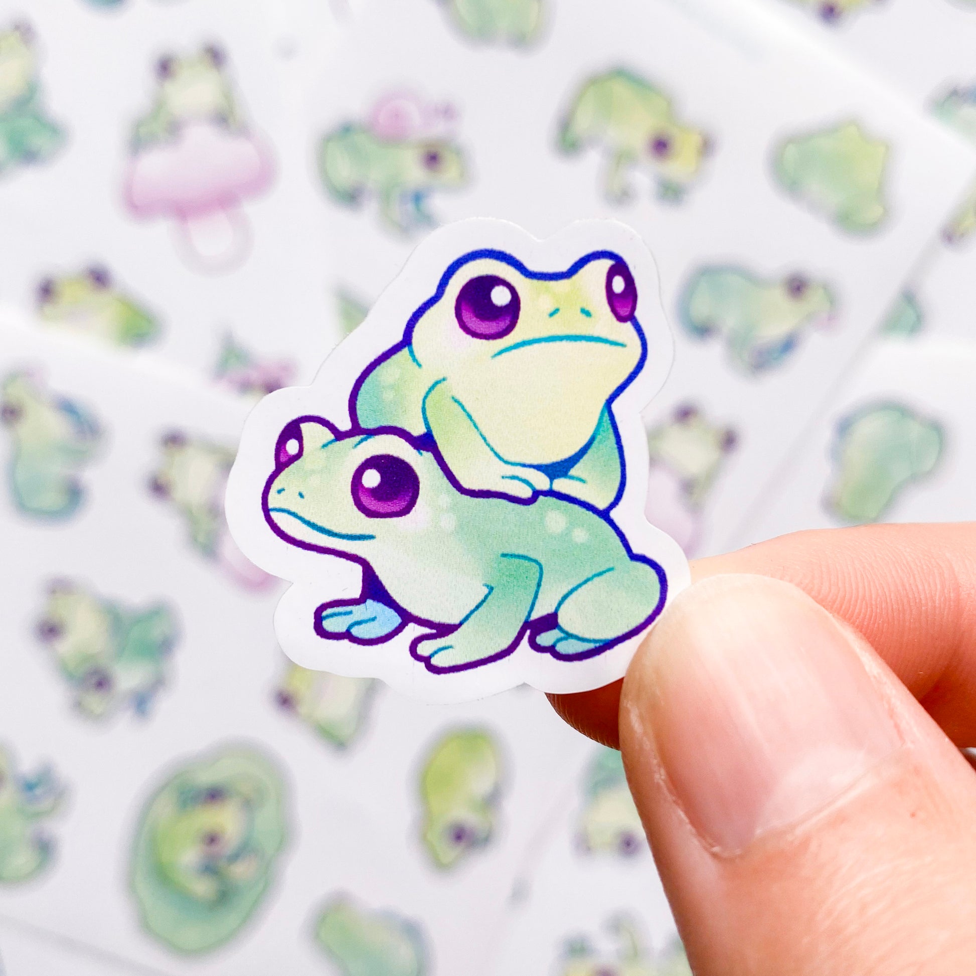 A sticker of two frogs sitting on top of each other. Each sticker is an individual unique drawing of a frog in a different pose. Some poses are funny and very charming, like this one. The sticker sheet also includes one sitting on a mushroom, one with a snail on its back, one sticking his tongue out and one with a flower on it's head.