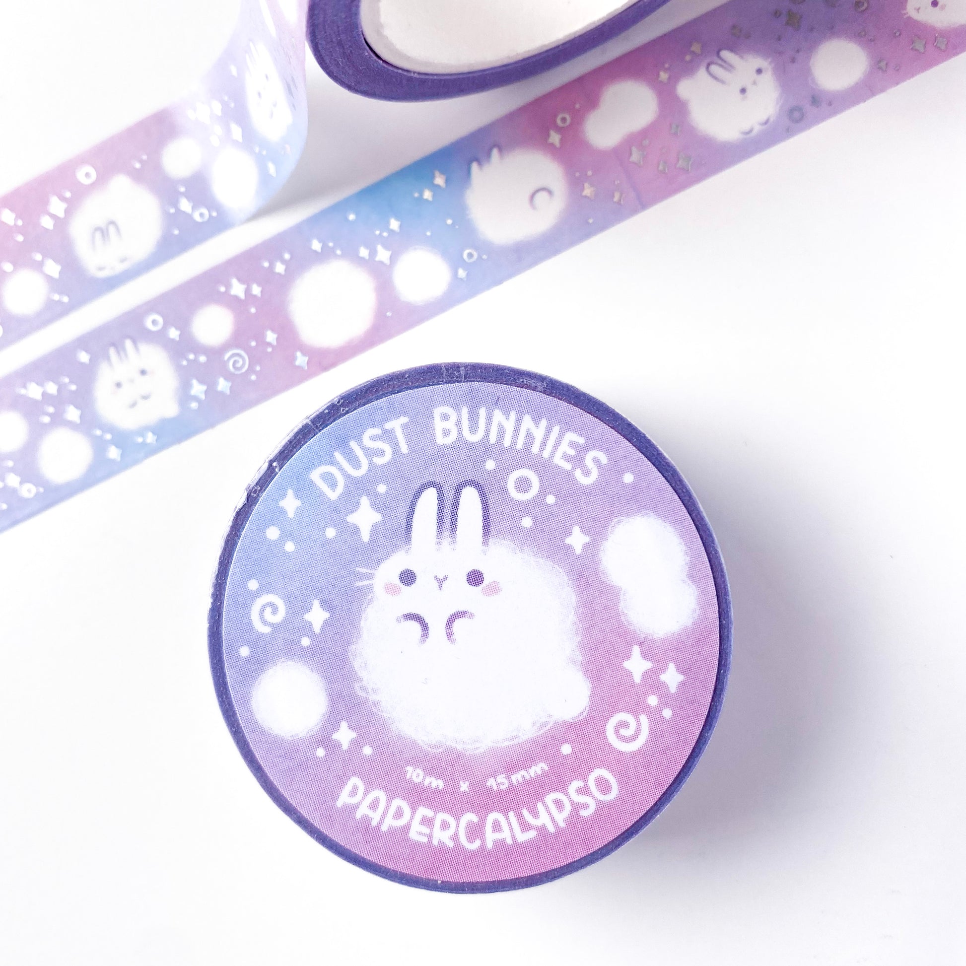 The roll of washi tape is packaged with shrink wrap plastic and a round sticker on one side. On it is an adorable fluffy bunny looking at us. It reads "Dust Bunnies, 10 metres by 15 milimetres, by Papercalypso"