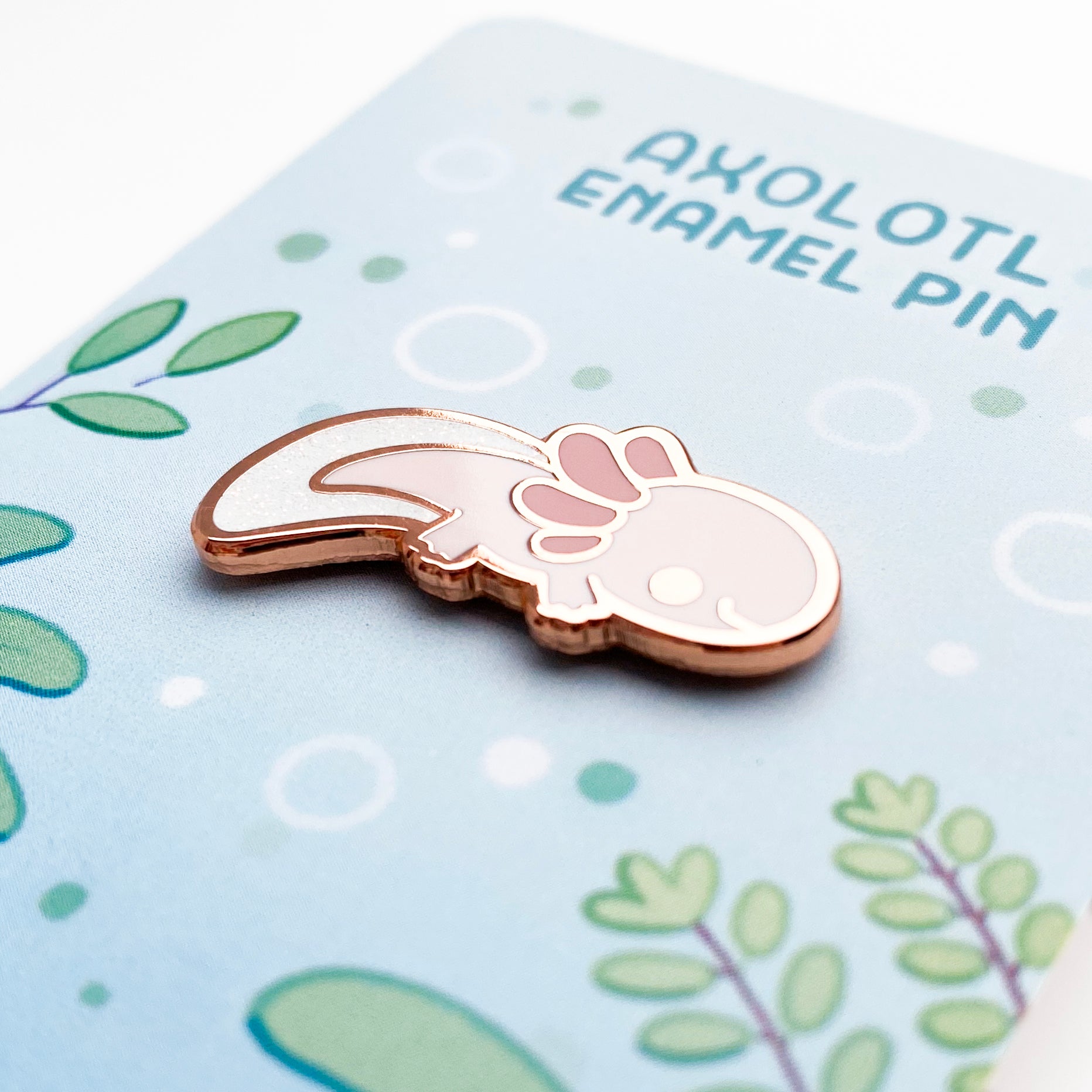 A close-up of the pin so you can see the details. The glitter accents sparkle in the light. The pin backing is an underwater freshwater lake with the title "Axolotl Enamel Pin".