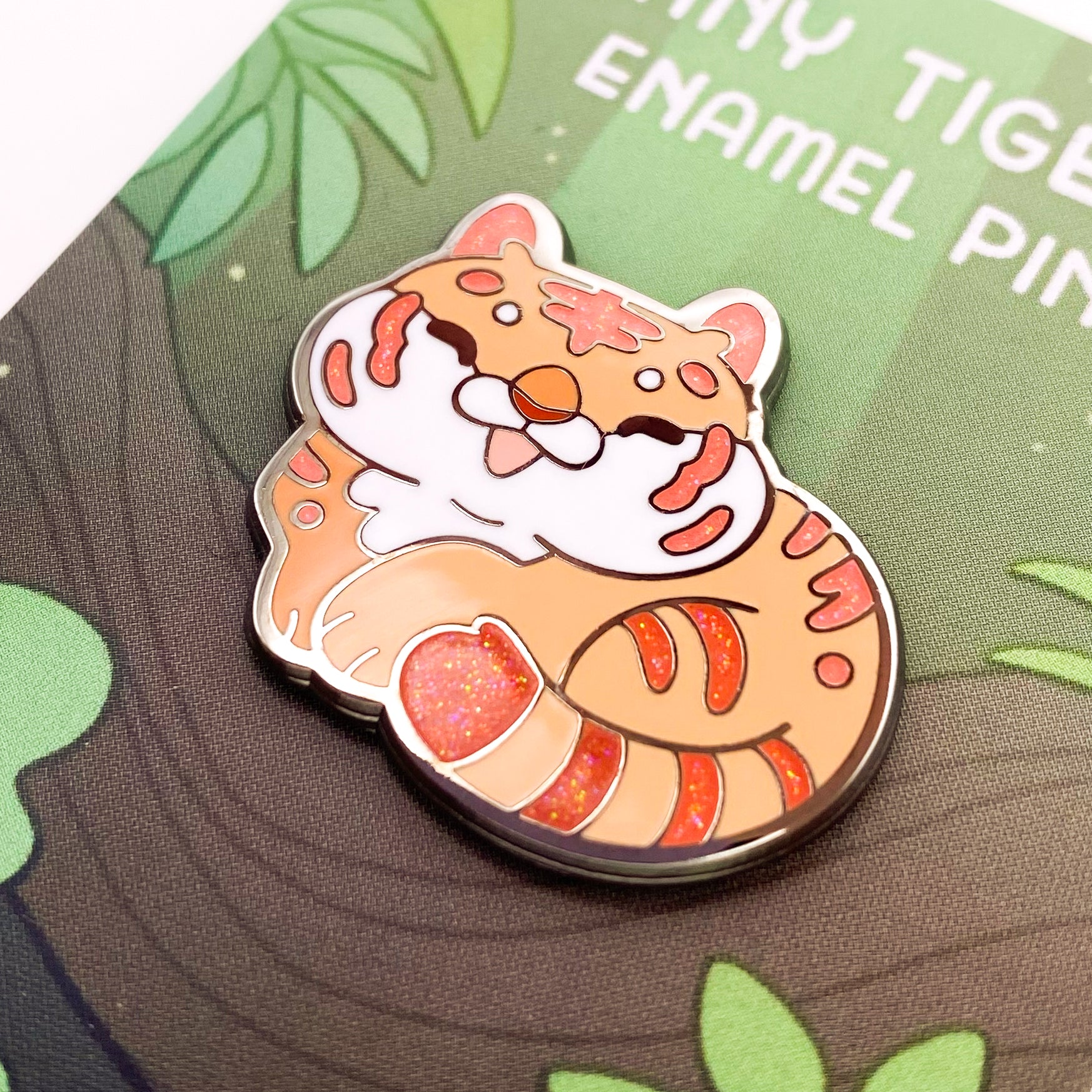 A close-up of the pin. The glitter accents sparkle in the light. The pin backing is a green jungle with the title "Tiny Tiger Enamel Pin."
