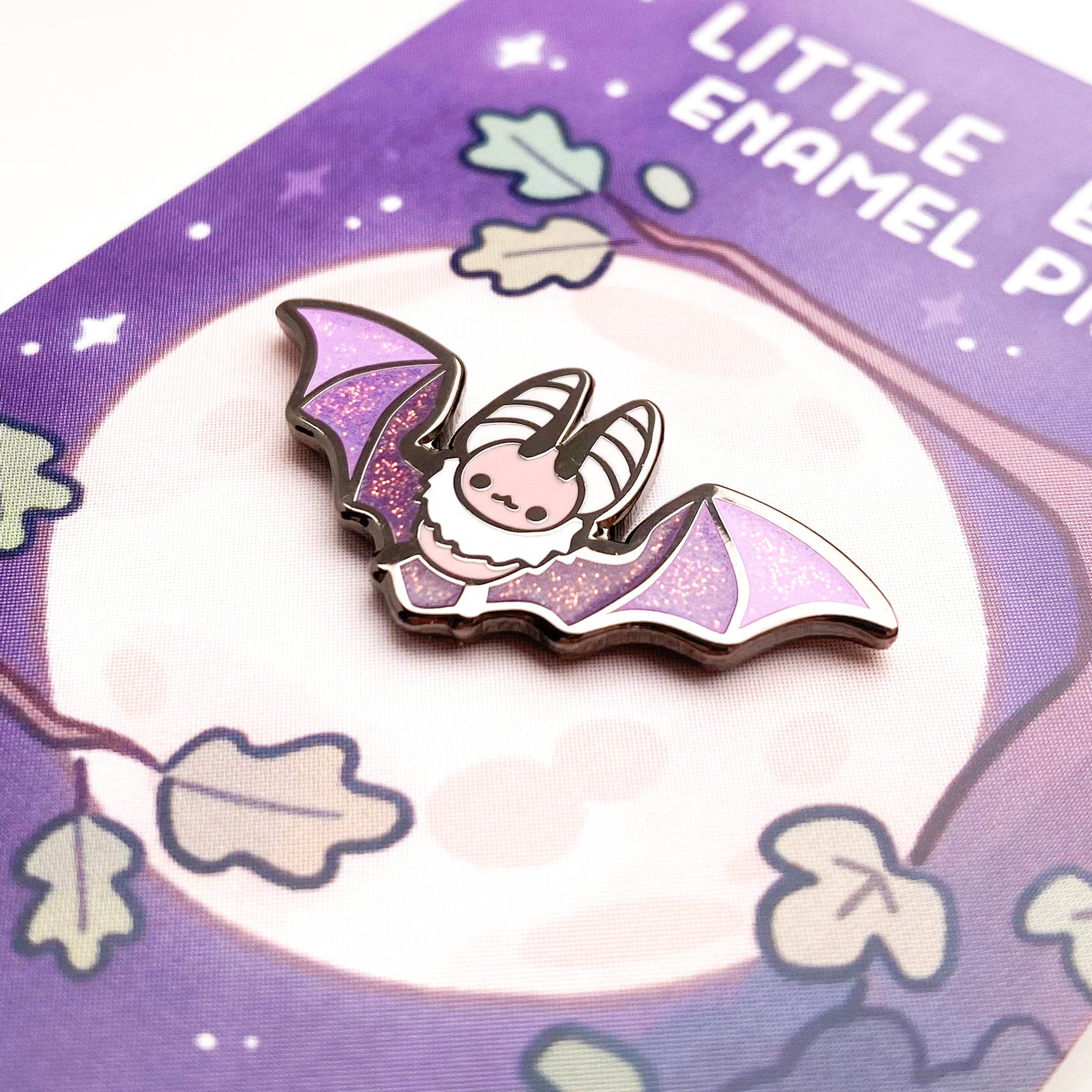 A close-up of the pin so you can see the details. The glitter accents sparkle in the light. The pin backing is purple nighttime oak forest scene with a full moon. The title reads "Little Bat Enamel Pin".