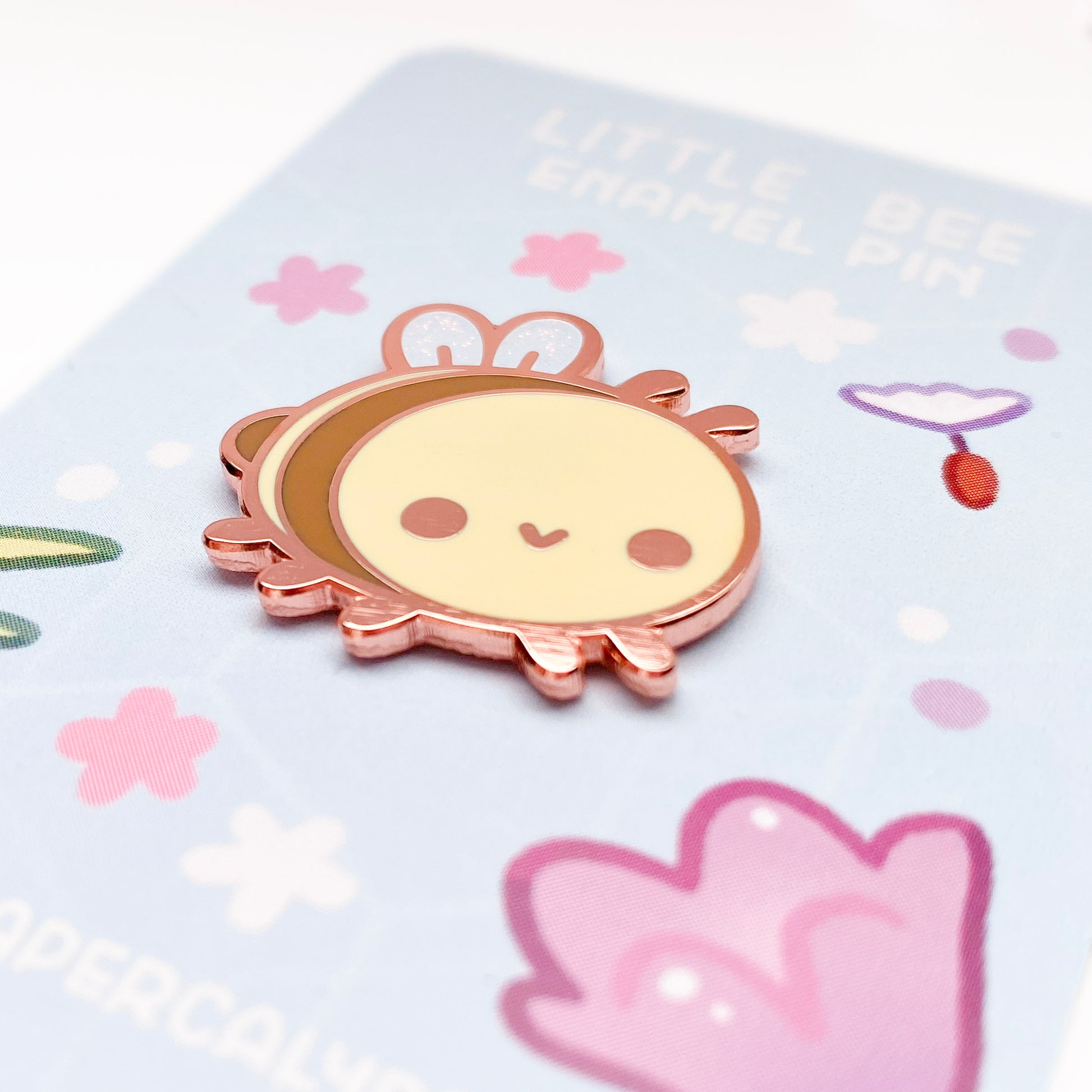 A close-up of the pin so you can see the details. The glitter accents on the wings sparkle in the light. The pin backing is a purple flower garden with the title "Little Bee Enamel Pin".