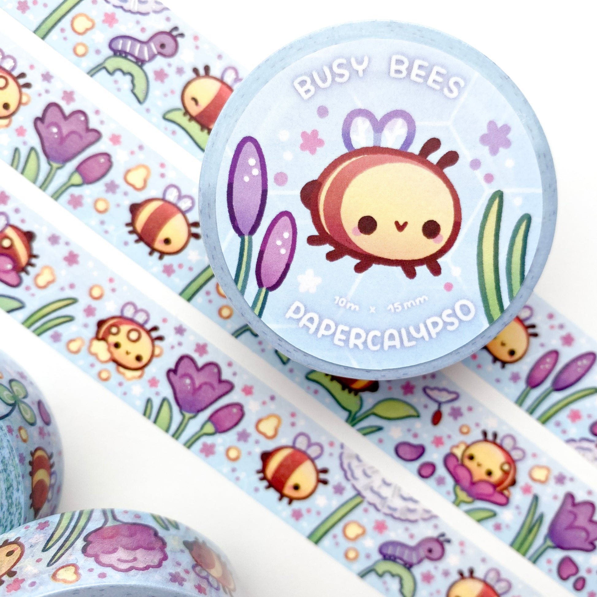 kawaii bee themed washi tape featuring pastel round bees flying in a spring meadow with a blue sky and plants. The bees are happily working and collecting pollen among the crocus flowers, dandelions and clovers. One bee is saying hi to a caterpillar!