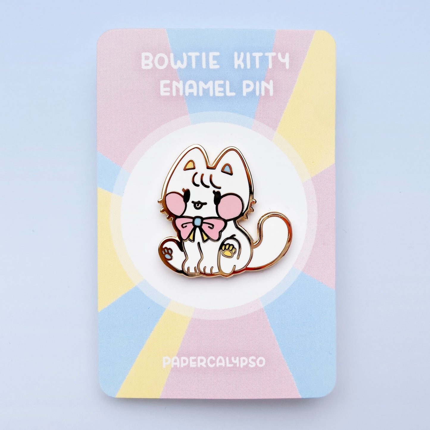 A gold metal enamel pin of a white cat with a cute bow around its neck. It's sitting down and sticking out its tongue. It has big pink blush spots, and pink blue and yello padpads and bow. Its proportions are round and stubby, making it appear cuter.