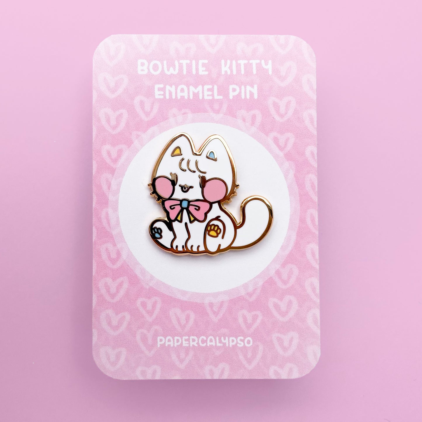 A gold metal enamel pin of a white cat with a cute bow around its neck. It's sitting down and sticking out its tongue. It has big pink blush spots and pink blue and yellow colours in its pawpads and bow. Its proportions are round and stubby, making it appear cuter.