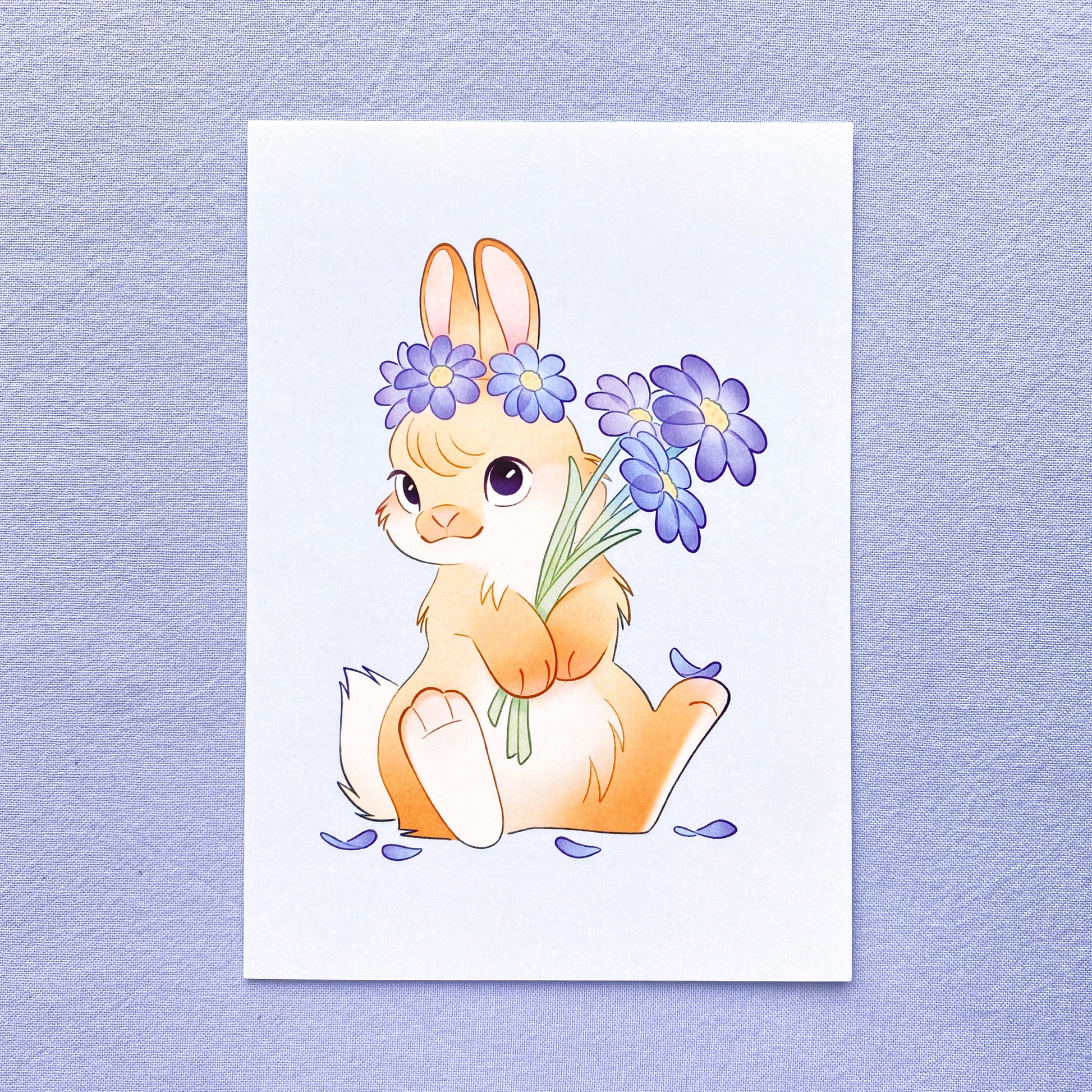 the card is a drawing of an domesticated pet bunny rabbit holding a bouquet of blue flowers while sitting. On its head is a flower crown and around it are petals. The background is a light blue, there is no text on the card so it has many uses.