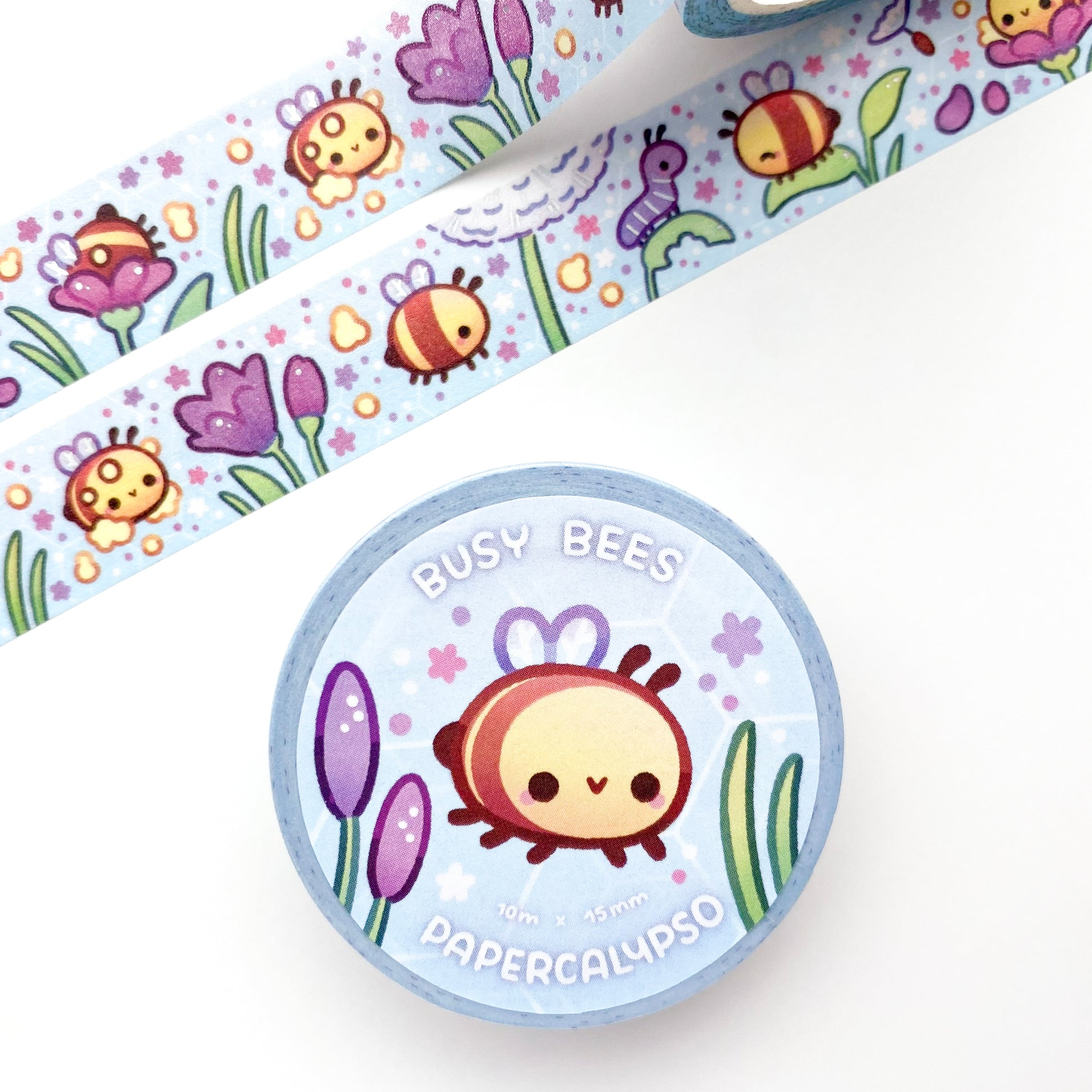the roll of washi tape is packaged with shrink wrap plastic and a round sticker on one side. On it is an adorable happy bee surrounded by flowers and other plants. It reads "Busy Bees, 10 metres by 15 milimetres, by Papercalypso"