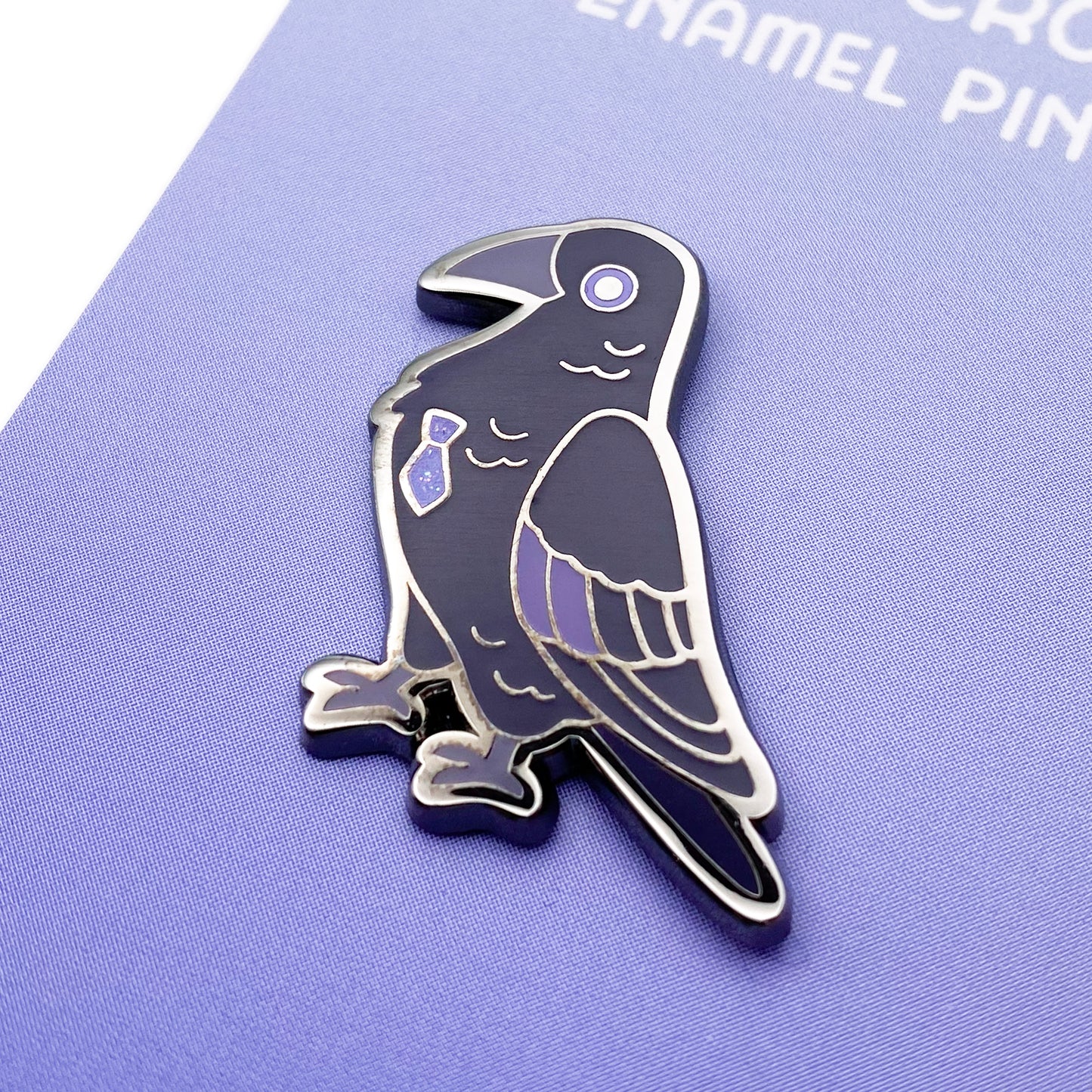 A close-up of the enamel pin so you can see the details. The glitter accents sparkle in the light. The pin backing is a purple gradient with the title "Fancy Crow Enamel Pin."