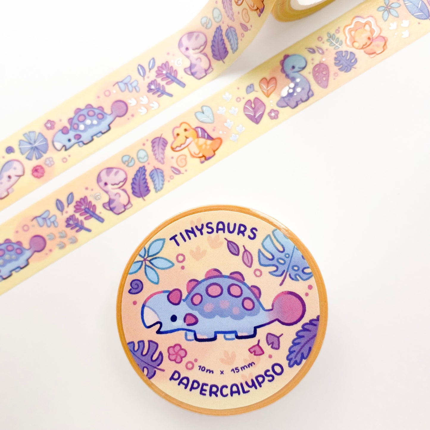 the roll of washi tape is packaged with shrink wrap plastic and a round sticker on one side. On it is a cute blue ankylosaurus surrounded by different plants. It reads "Tinysaurs, 10 metres by 15 milimetres, by Papercalypso" 