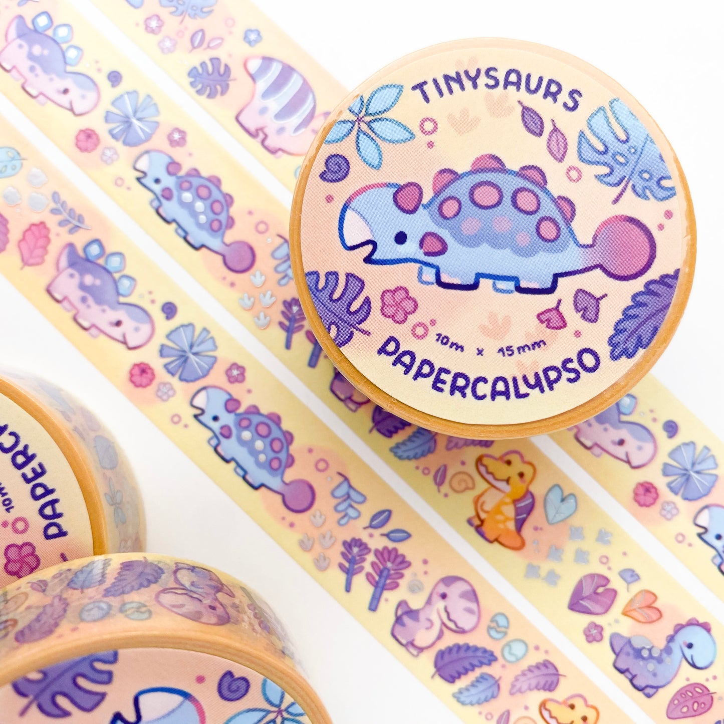 kawaii paleo themed washi tape featuring cartoon dinos on a yellow background with tropical plants. Featured dinosaurs are: Tyrannosaurus Rex, Ankylosaurus,  Triceratops, Stegosaurus, Brachiosaurus, Spinosaurus, Hadrosaurus and Pterosaurs.