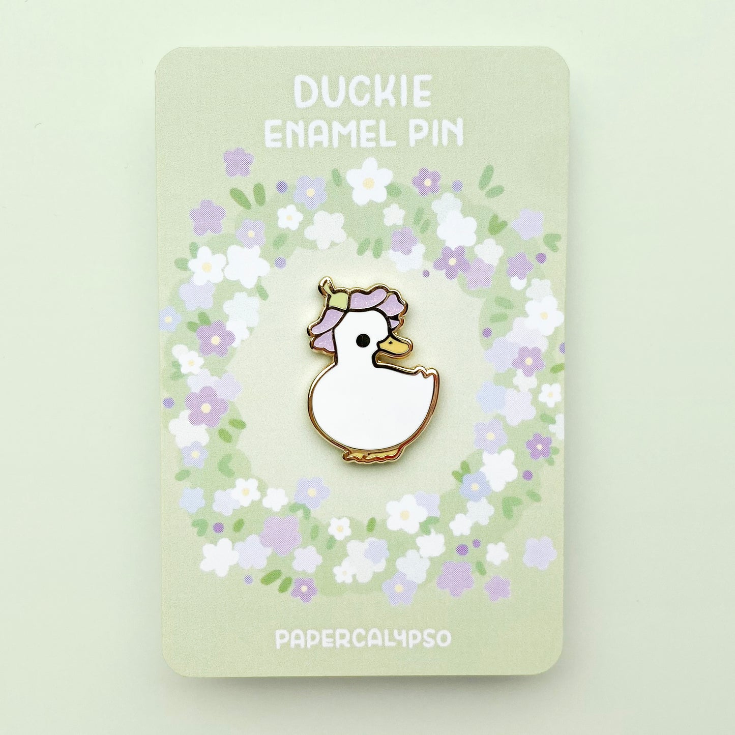 A gold plated hard enamel pin of a domestic pet white duck wearing a purple glittery flower as a hat.