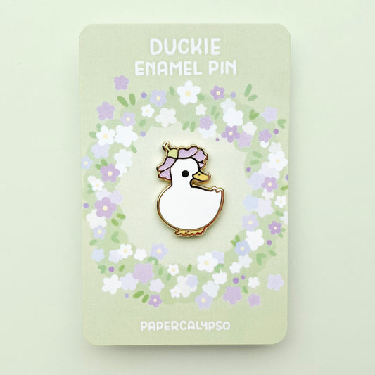 A gold plated hard enamel pin of a domestic pet white duck wearing a purple glittery flower as a hat.