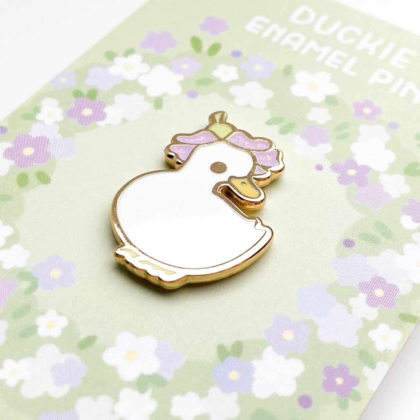 A close-up of the pin so you can see the details. The glitter accents sparkle in the light. The pin backing is a ring of flowers with the title "Duckie Enamel Pin."