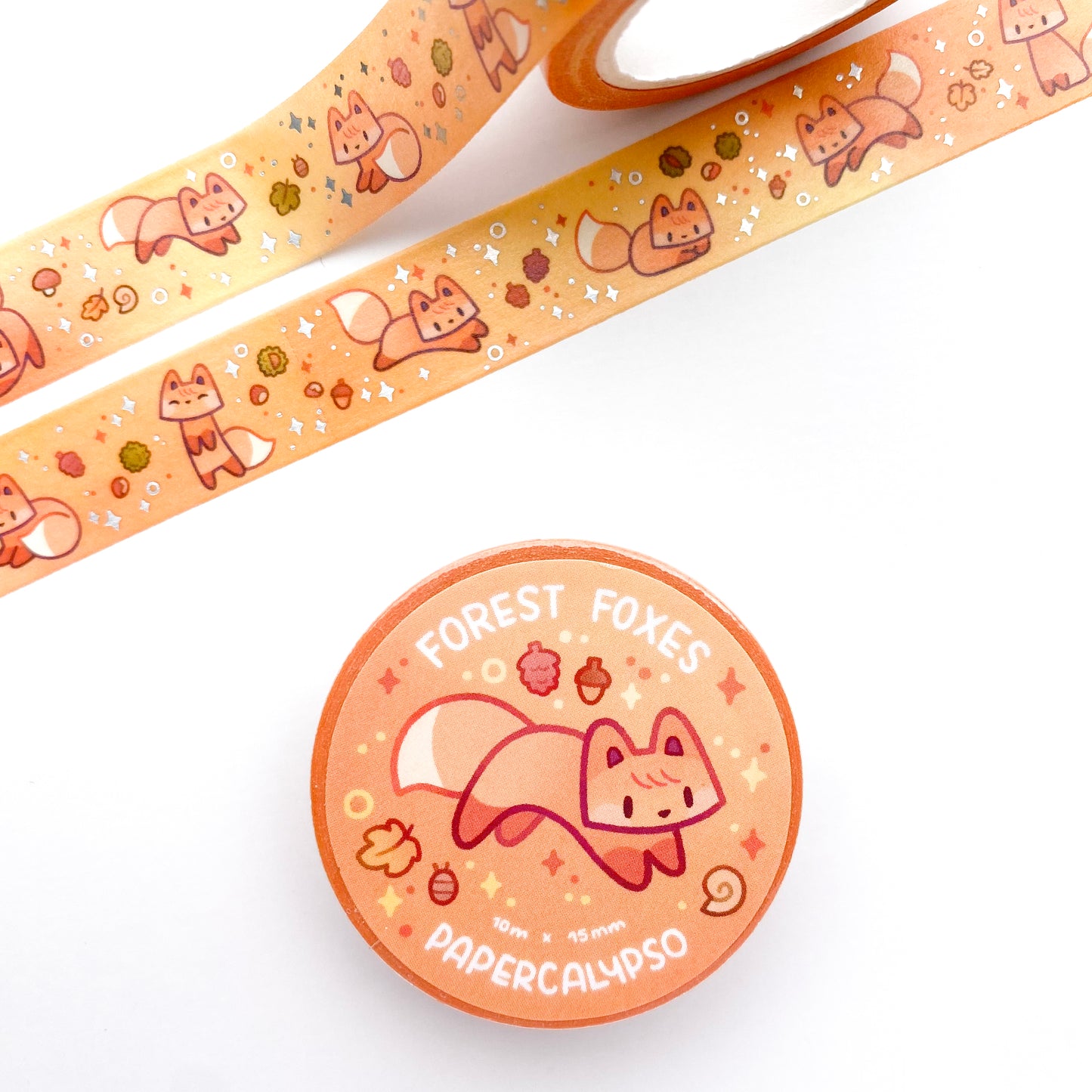 the roll of washi tape is packaged with shrink wrap plastic and a round sticker on one side. On it is an adorable playful fox along with a pill bug, a snail shell, and a maple tree leaf. It reads "Forest Foxes, 10 metres by 15 milimetres, by Papercalypso"