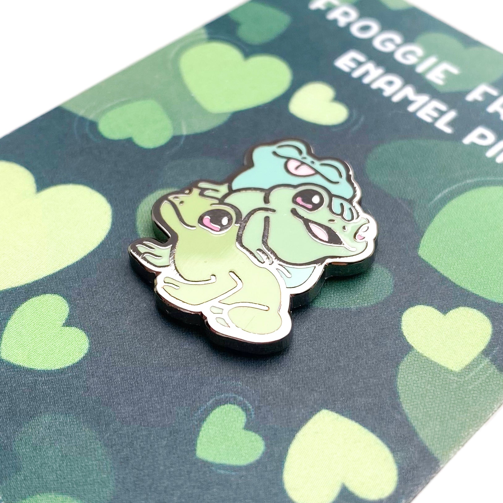 A close-up of the pin so you can see the details. The pin backing is a green lilly pad pond with the title "Froggie Friends Enamel Pin".