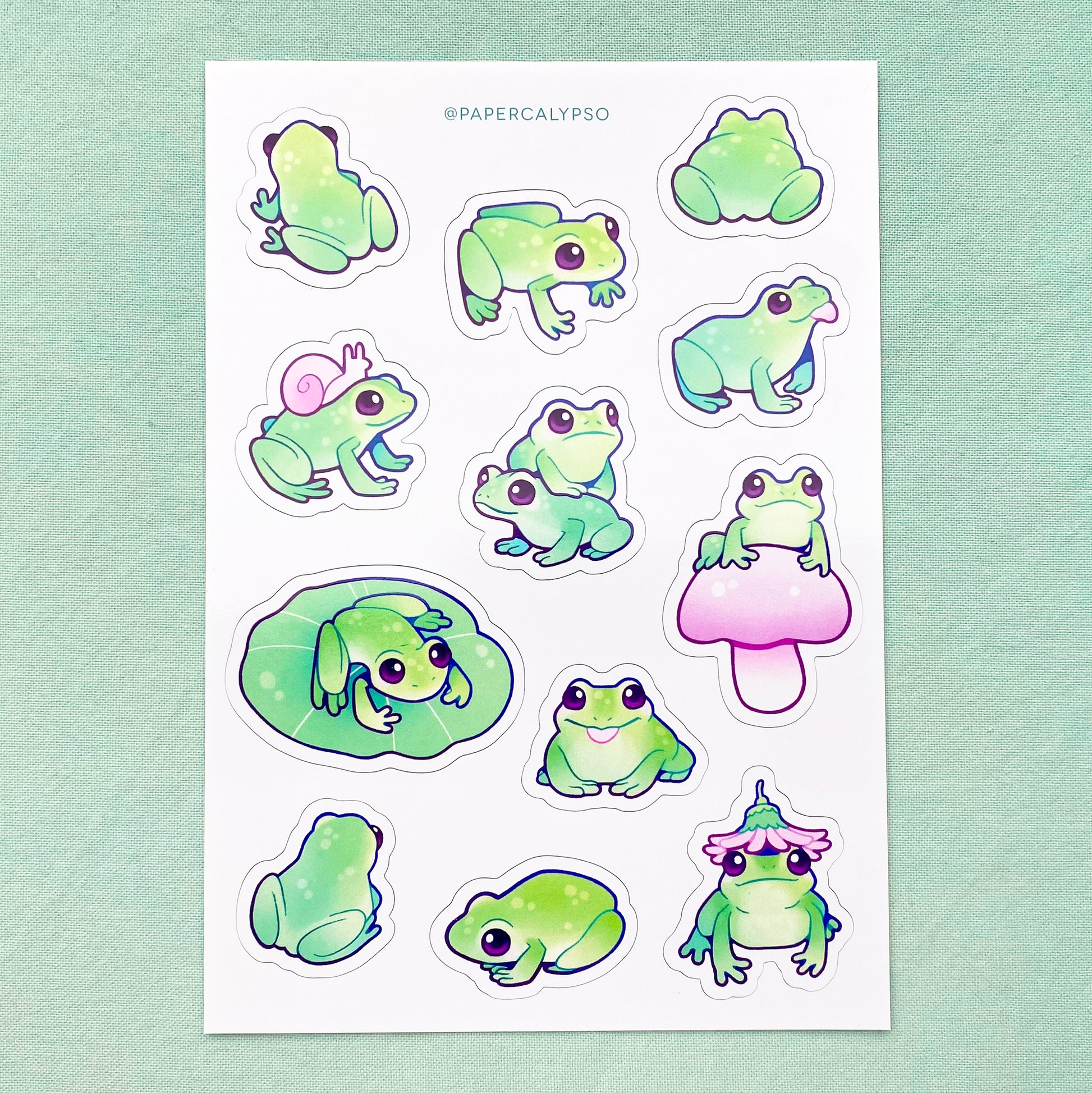 The frogs are green with prominent blue line art. They have rounded cartoon shapes, so they appear cuter. Their eyes are big and purple. The sticker sheet has 12 unique stickers printed on white matte thin vinyl. This makes them sturdy and waterproof, but not too thick to use in a journal.