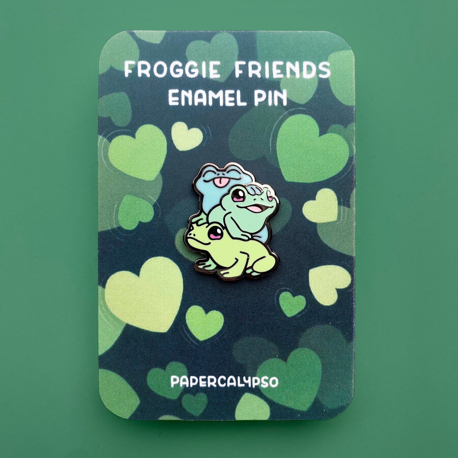 A black nickel hard enamel pin of three froggies sitting on top of each other. They're all happily smiling as if they're posing for a picture together. They're each a different shade of green and their eyes are pink.