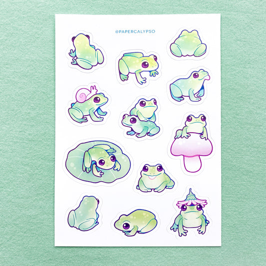 The frogs are green with prominent blue line art. They have rounded cartoon shapes, so they appear cuter. Their eyes are big and purple. The sticker sheet has 12 unique stickers printed on white matte thin vinyl. This makes them sturdy and waterproof, but not too thick to use in a journal.