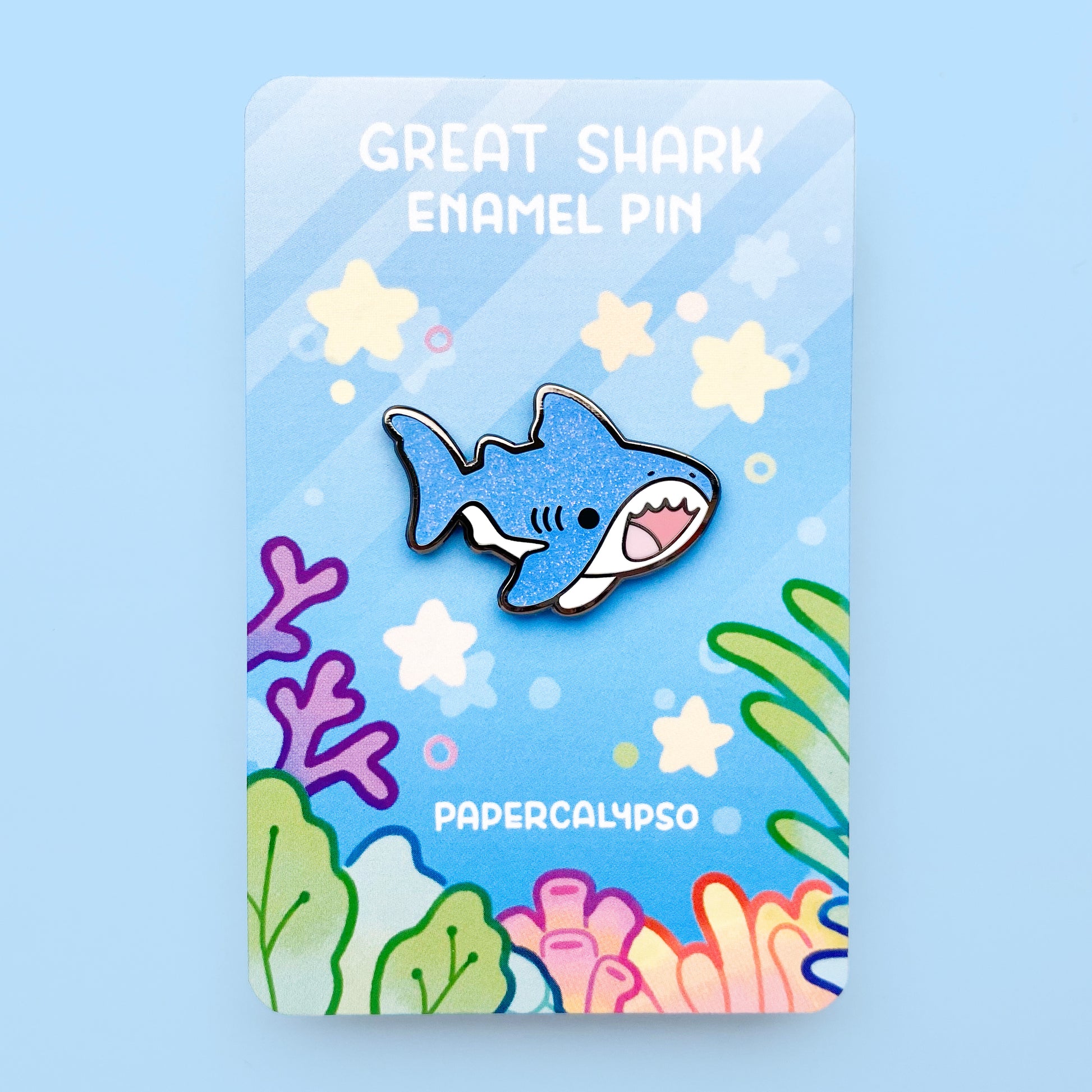 A black nickel hard enamel pin of a great white shark. He has a white tummy and glittery blue back. He's smiling with his mouth open and looks very cute!