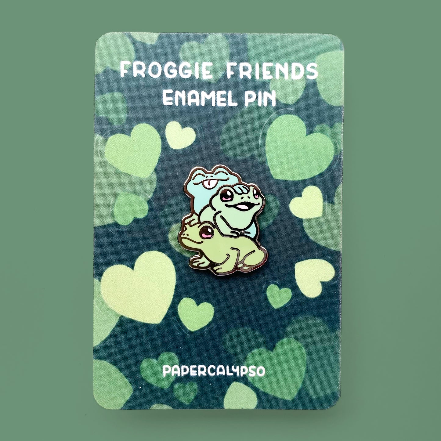 A black nickel hard enamel pin of three froggies sitting on top of each other. They're all happily smiling as if they're posing for a picture together. They're each a different shade of green and their eyes are pink.