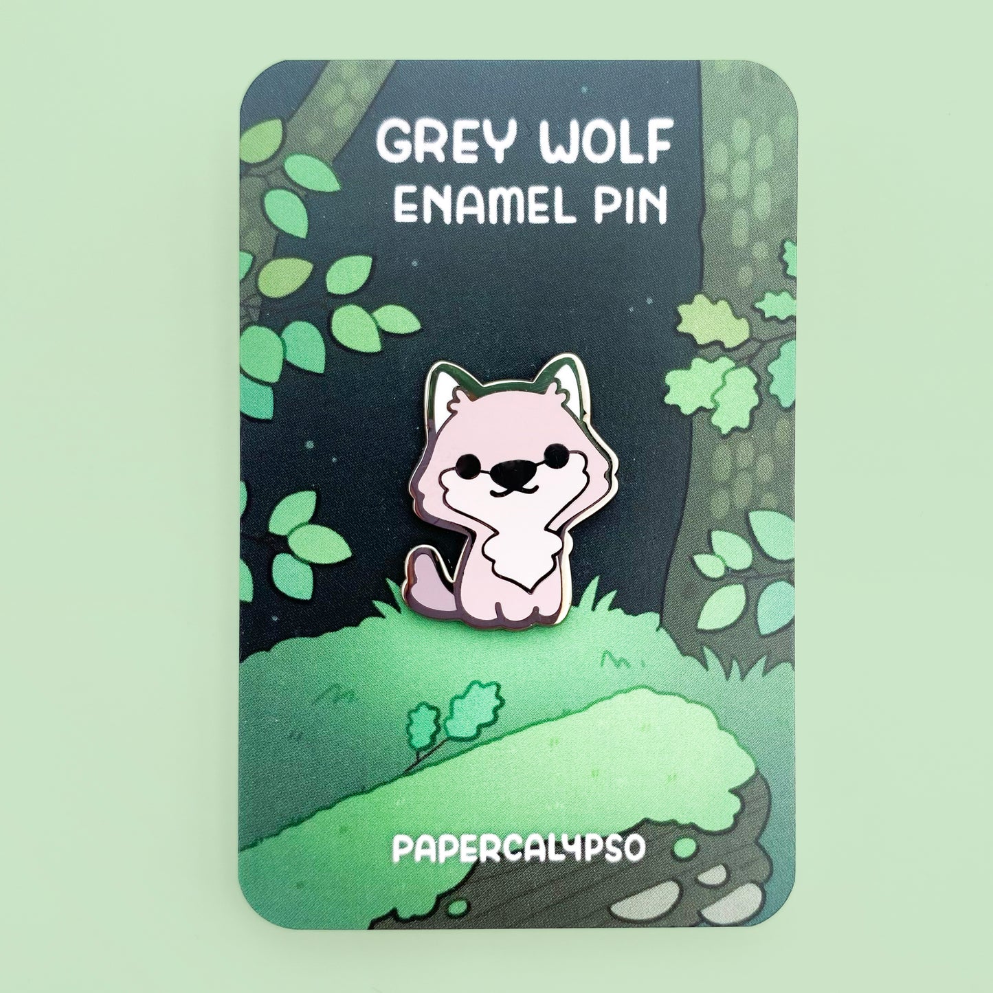 A nickel plated hard enamel pin of a cute wild grey wolf sitting and smiling. The backing card shows a green european forest.
