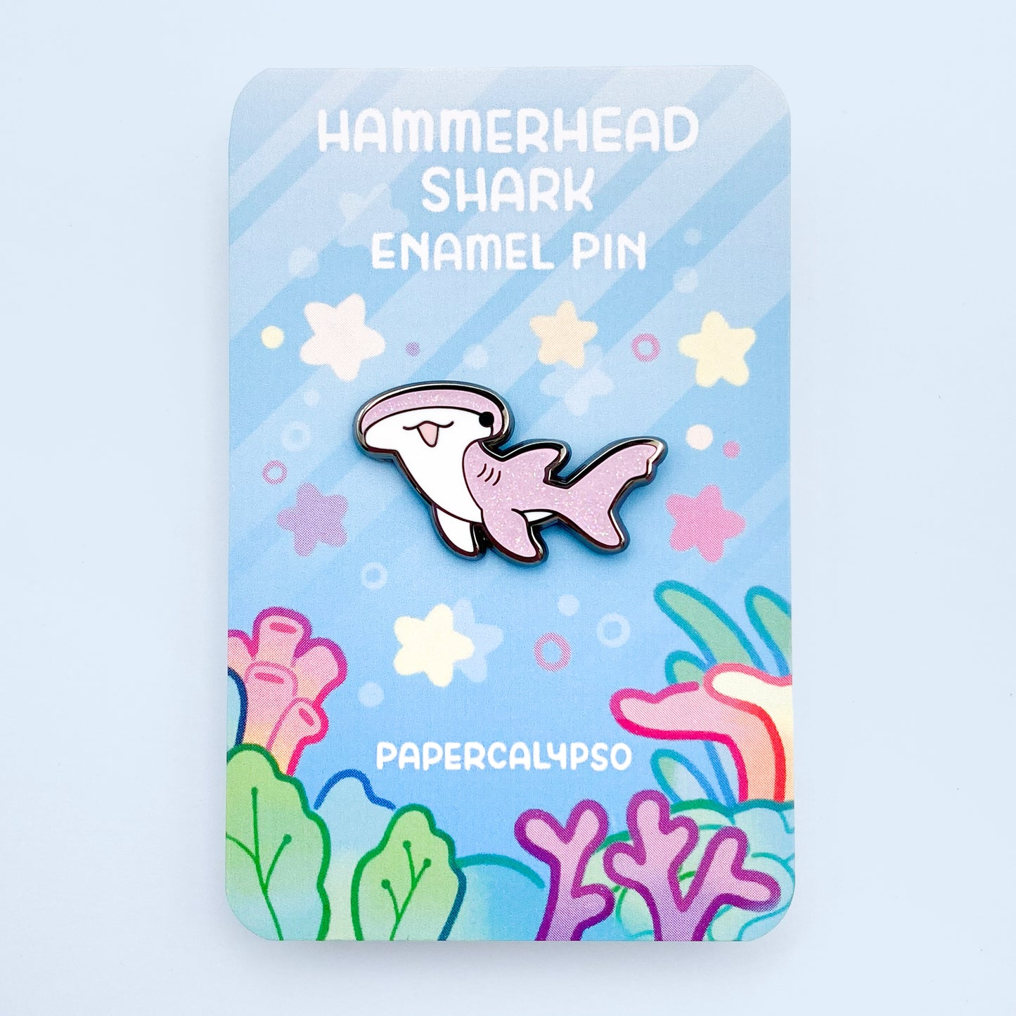 A black nickel hard enamel pin of a hammerhead shark. He has a white tummy and glittery pink back. He's smiling with his mouth open and looks very cute!