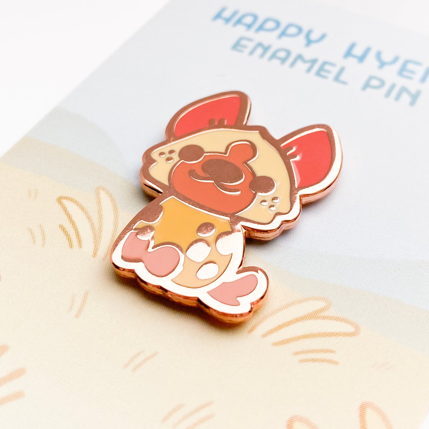 A close-up of the pin so you can see the details. The pin backing is grasslands scene with the title "Happy Hyena Enamel Pin".