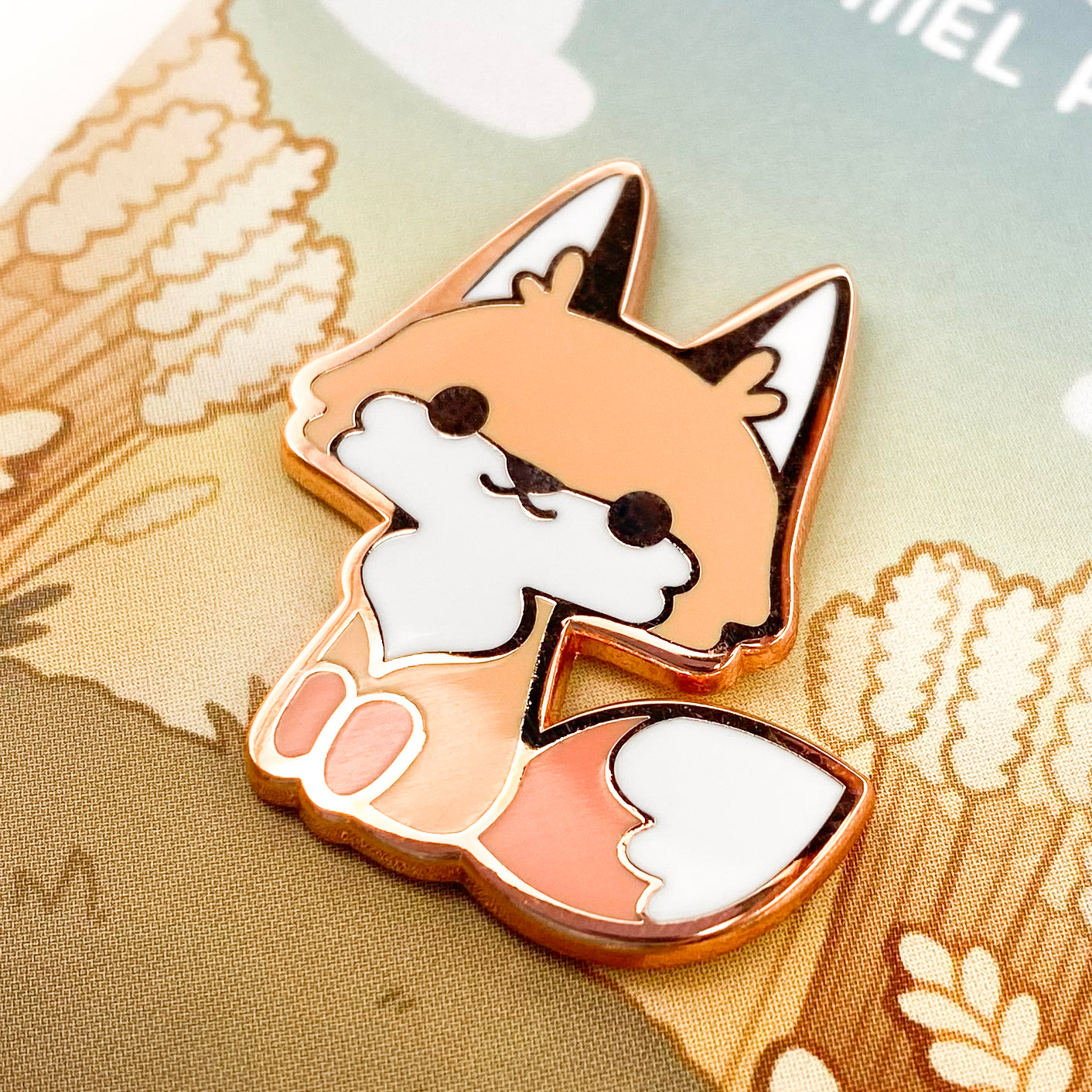 A close-up of the red fox pin. The pin backing is a wheat field in the sunset, parted by a stretch of grass, on which the fox is sitting. The title says "Red Fox Enamel Pin."