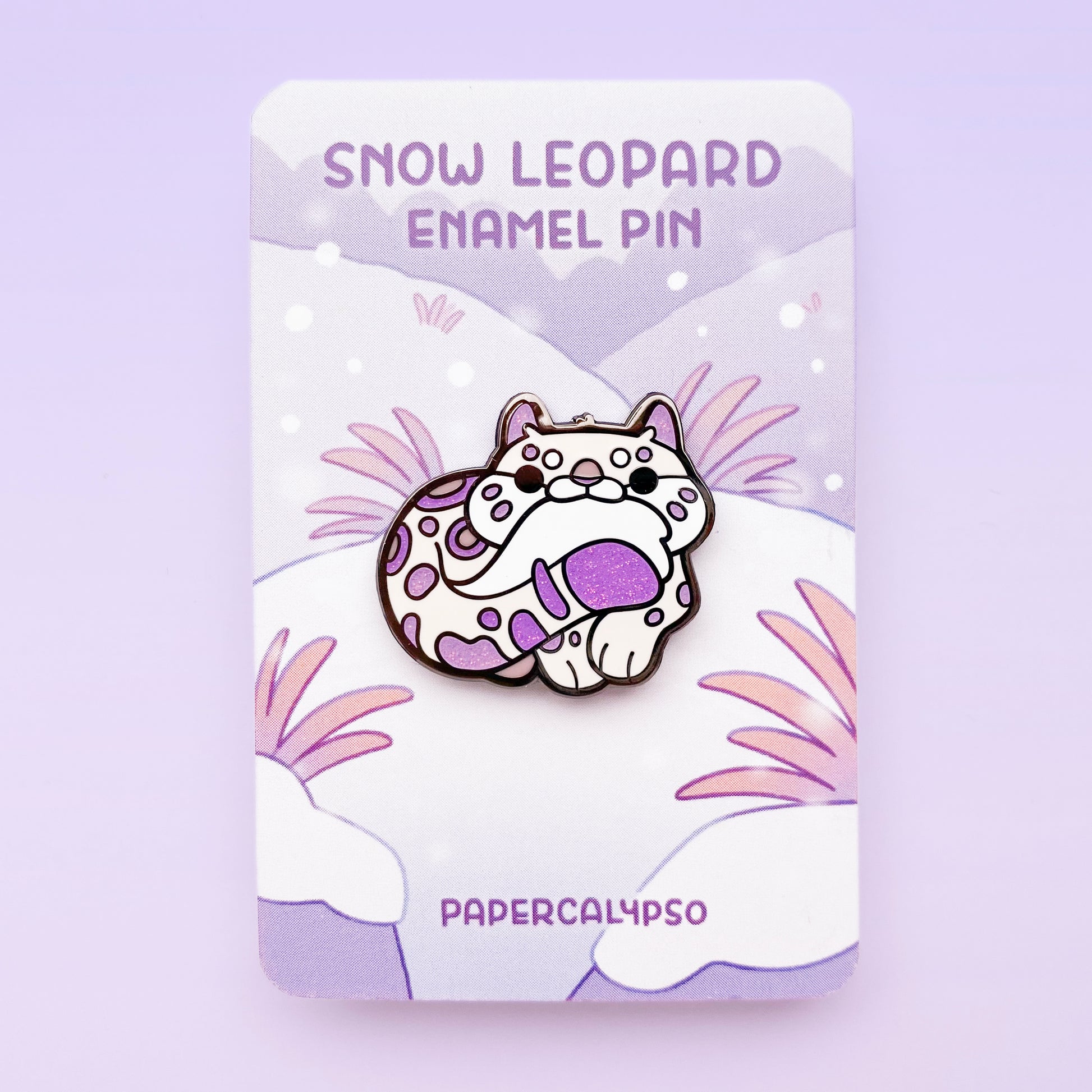 A black nickel hard enamel pin of a cute snow leopard biting its own tail. He's a cream colour with purple glitter spots.