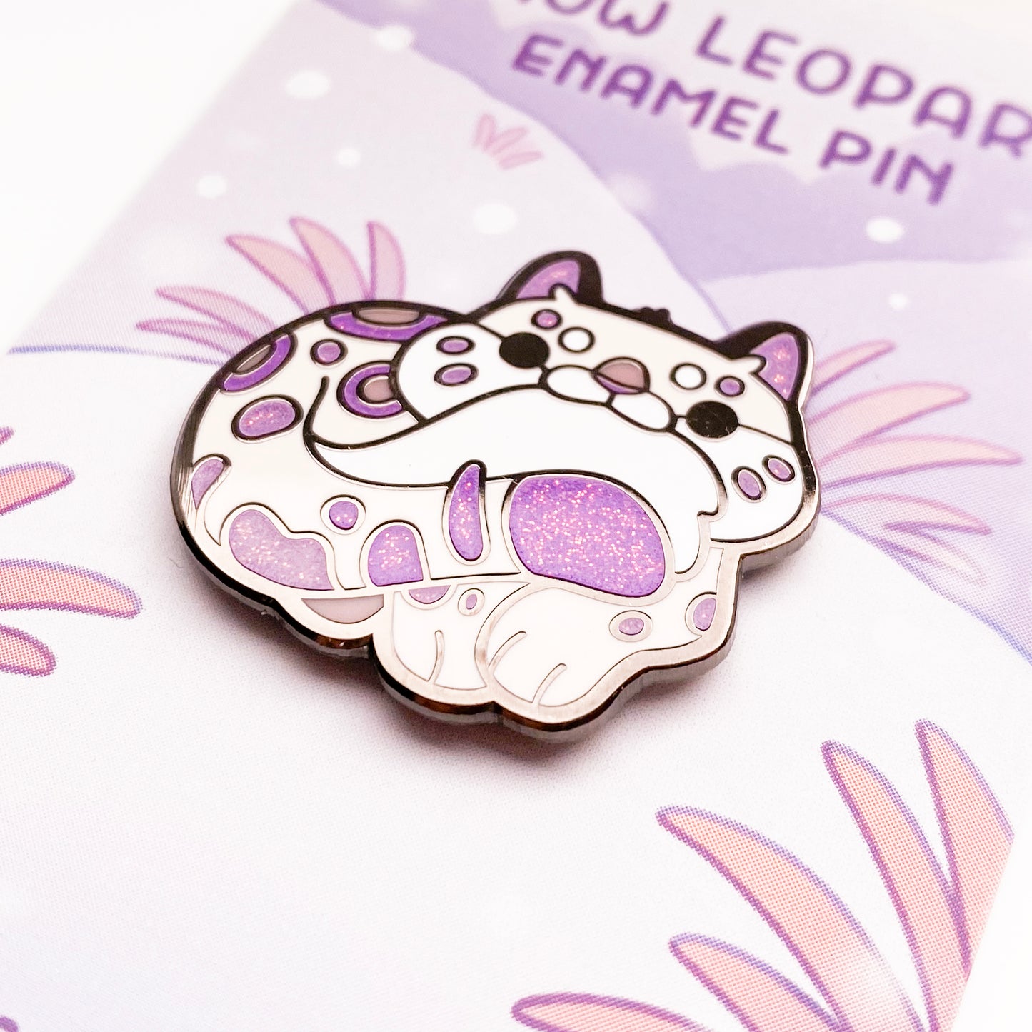 A close-up of the pin so you can see the details. The glitter accents sparkle in the light. The pin backing is a snowy mountain scene with the title "Snow Leopard Enamel Pin".