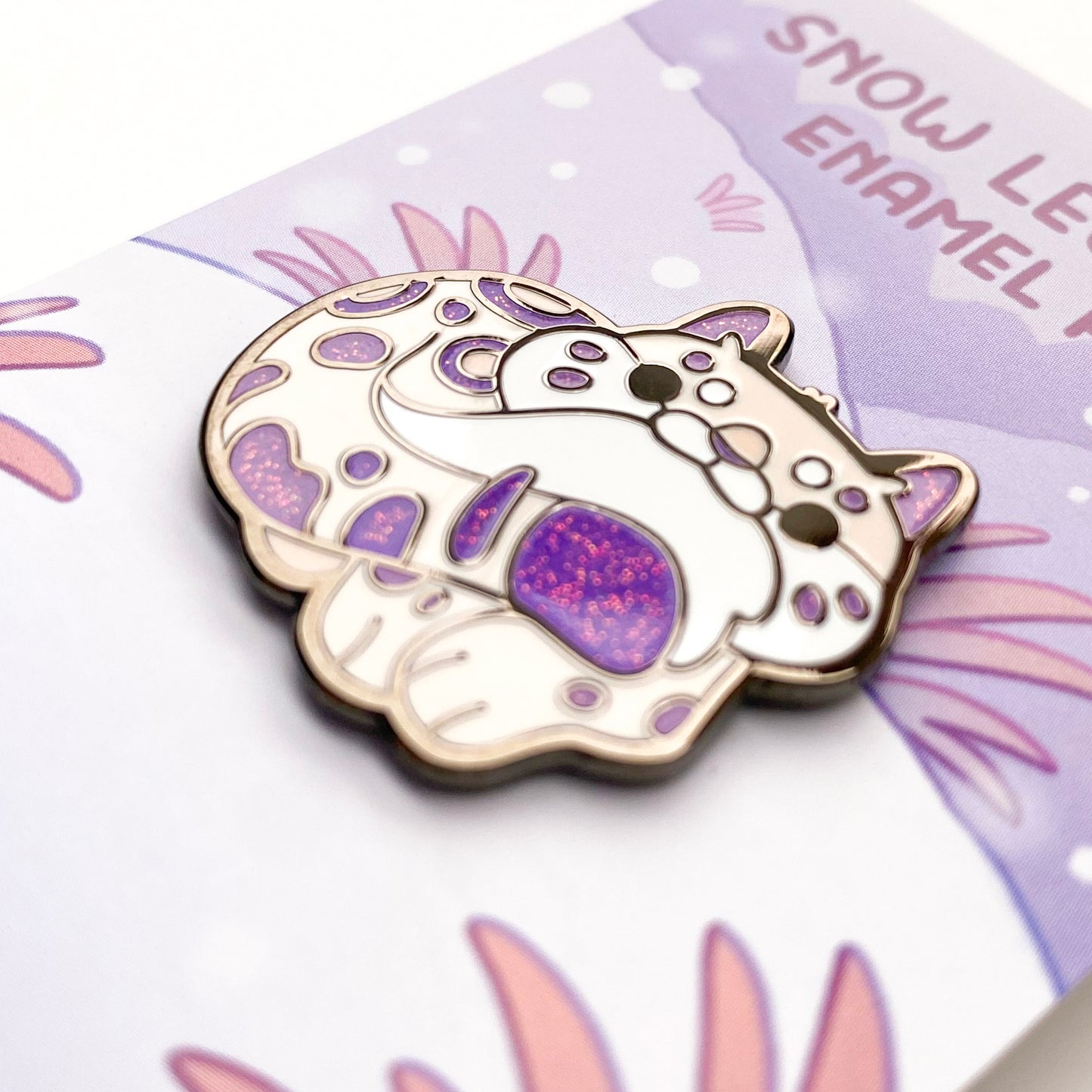 A close-up of the pin so you can see the details. The glitter accents sparkle in the light. The pin backing is a snowy mountain scene with the title "Snow Leopard Enamel Pin".
