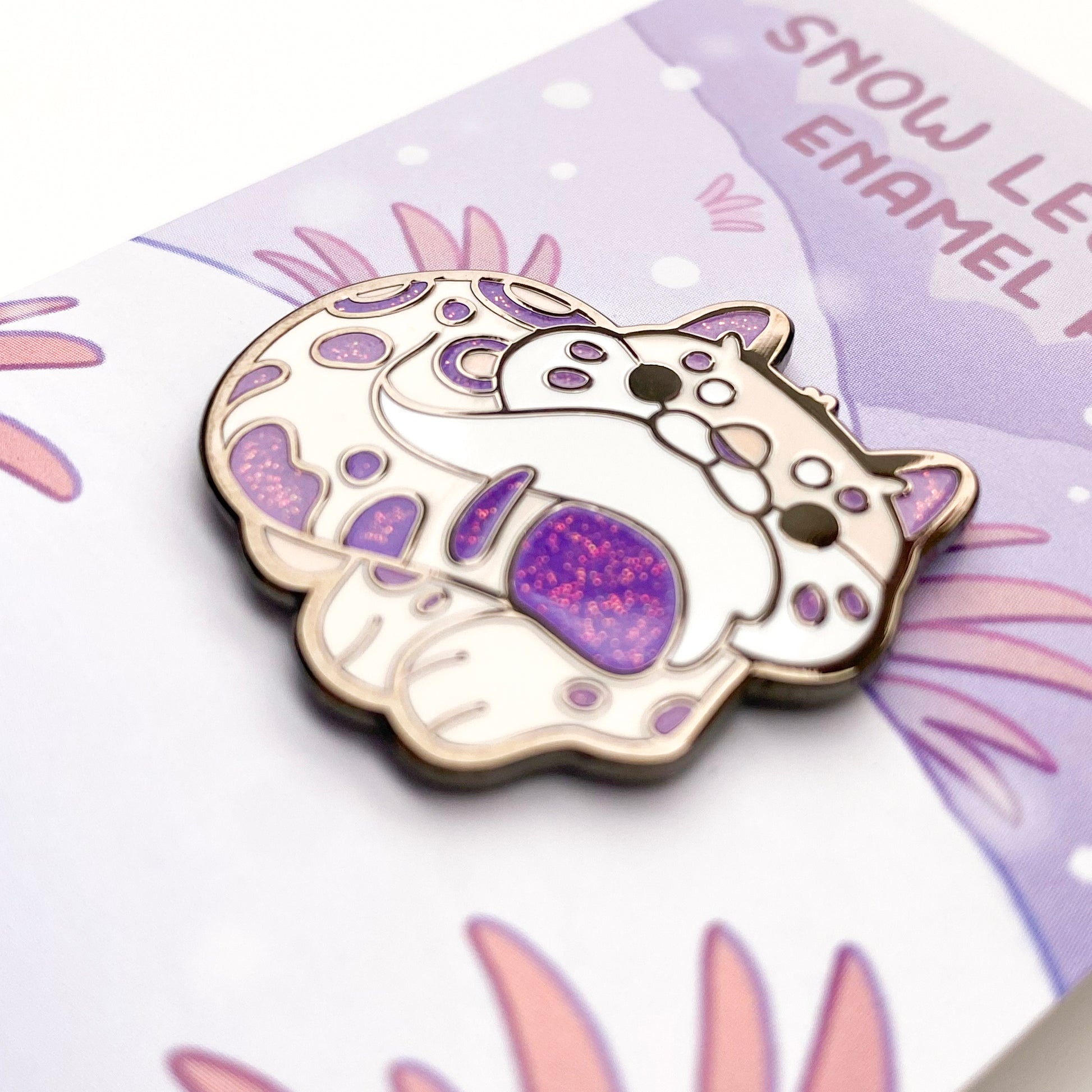 A close-up of the pin so you can see the details. The glitter accents sparkle in the light. The pin backing is a snowy mountain scene with the title "Snow Leopard Enamel Pin".