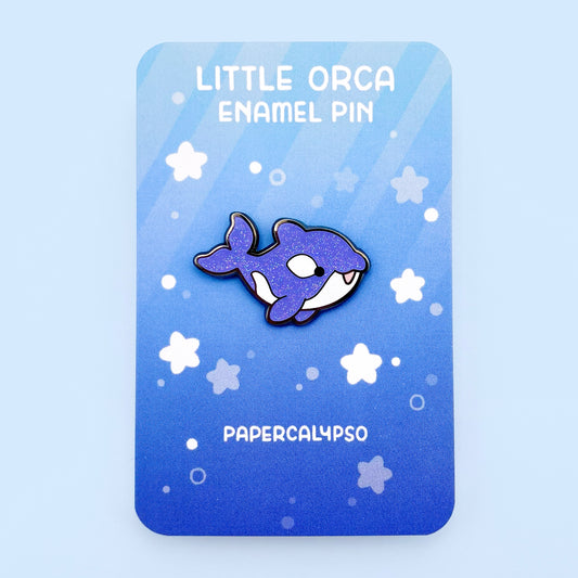 A black nickel plated hard enamel pin of an orca in the sea with a blue glittery back and white tummy.
