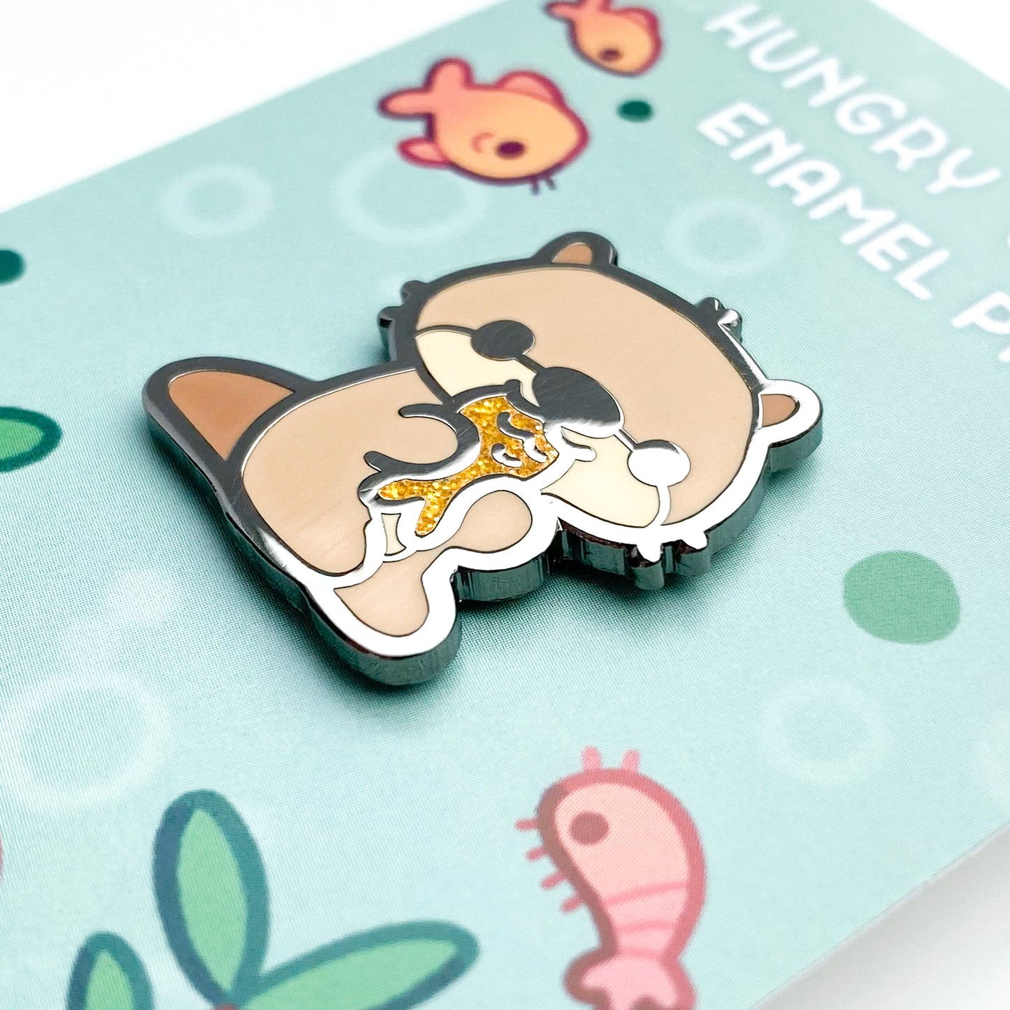 A close-up of the pin so you can see the details. The glitter accents sparkle in the light. The pin backing is an underwater scene with the title "Hungry Otter Enamel Pin".