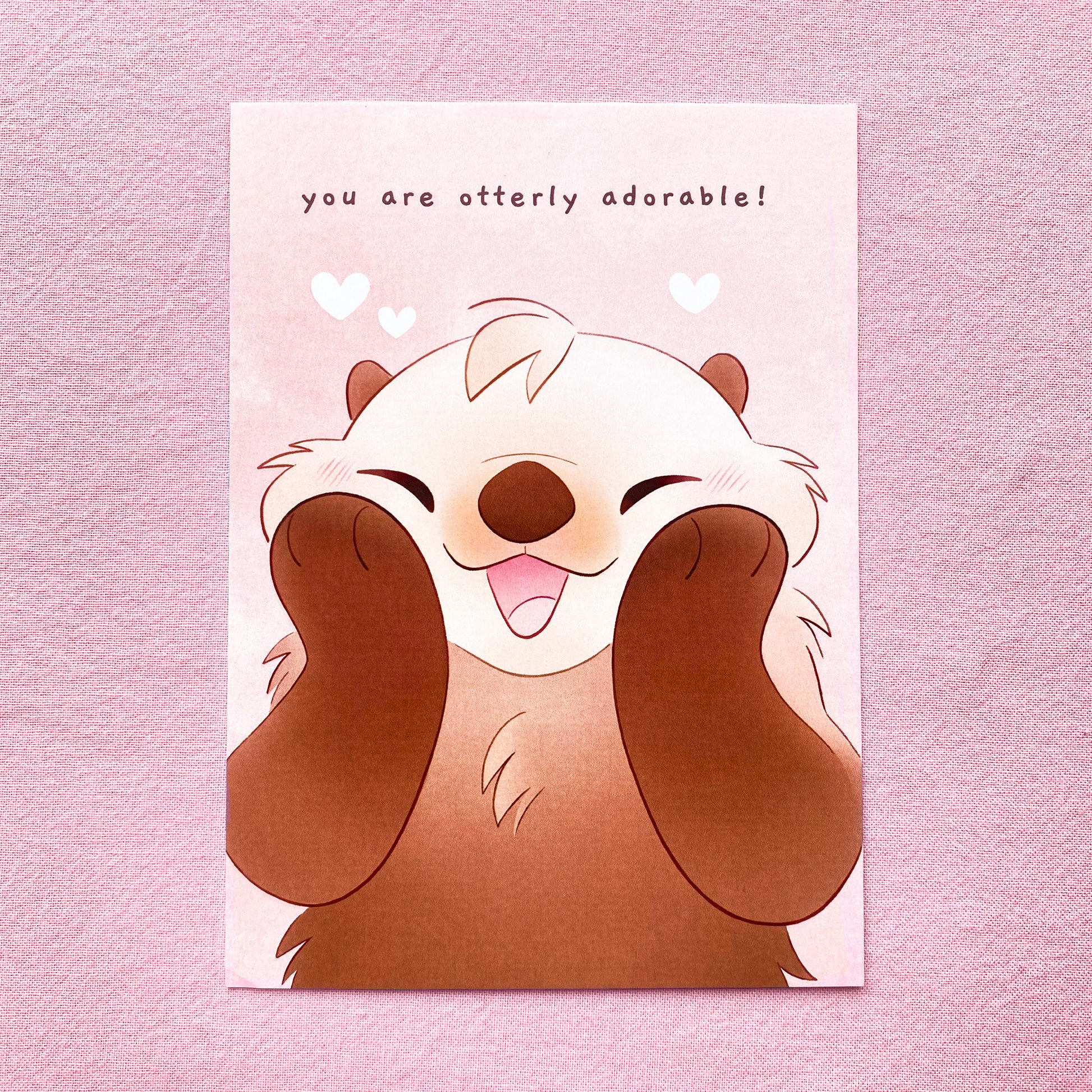 the card is a drawing of an american sea otter holding its white face and smiling with its mouth open. The background is pink with some white hearts. It's face and cheeks are squashed in a cute way. On the top is the text "You are otterly adorable"