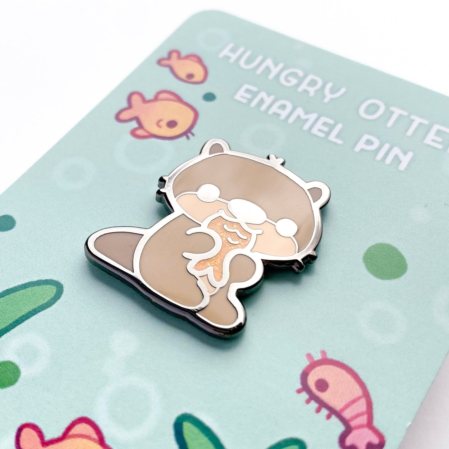 A close-up of the pin so you can see the details. The glitter accents sparkle in the light.  The pin backing features all the otter's underwater critter friends like fish, freshwater shrimp and a snail with some bubbles, plants and algae.