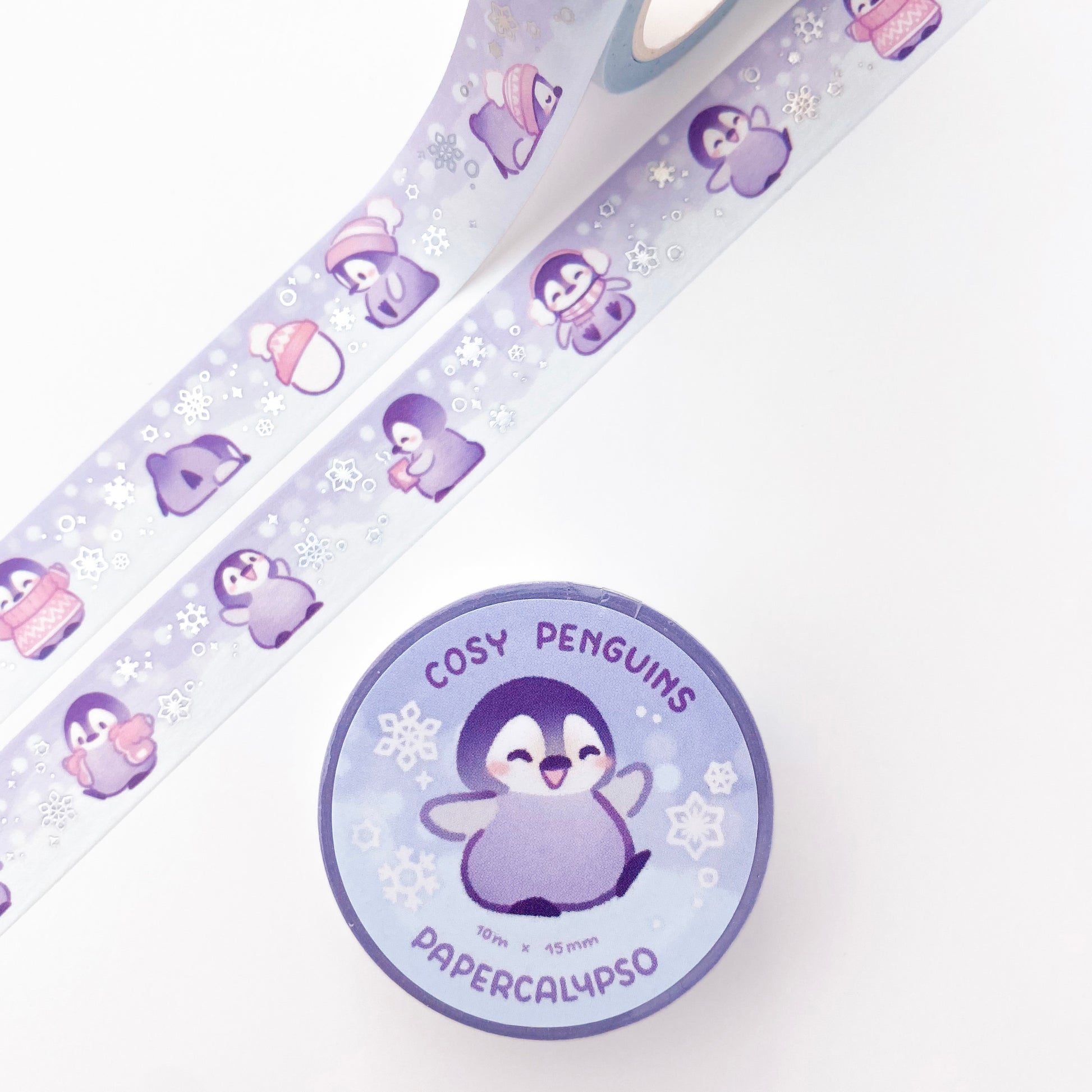 The roll of washi tape is packaged with shrink wrap plastic and a round sticker on one side. On it is an adorable baby emperor penguin chick lifting one of his feet. It reads "Cozy Penguins, 10 metres by 15 milimetres, by Papercalypso"