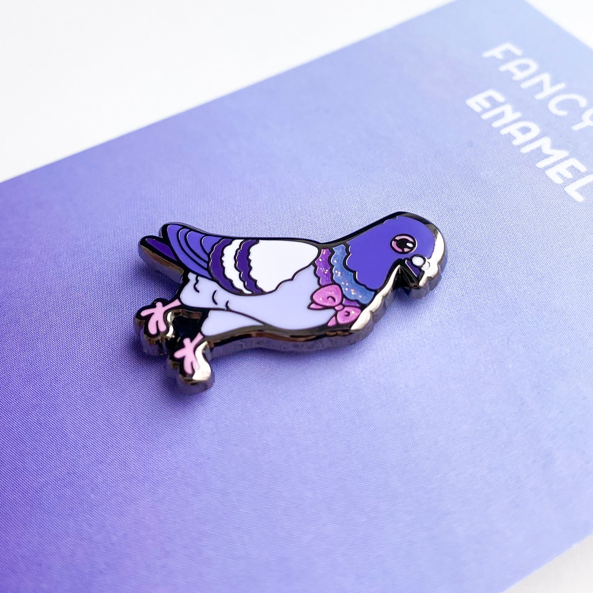 A close-up of the pin so you can see the details. The glitter accents sparkle in the light. The pin backing is a purple gradient with the title "Fancy Pigeon Enamel Pin".