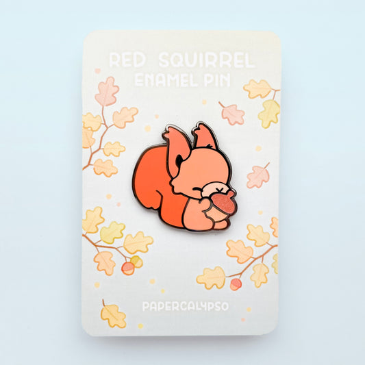 A black nickel hard enamel pin of a fluffy red squirrel eating a glittery brown acorn. He has a big fluffy tail, tufted ears and a little fringe. 