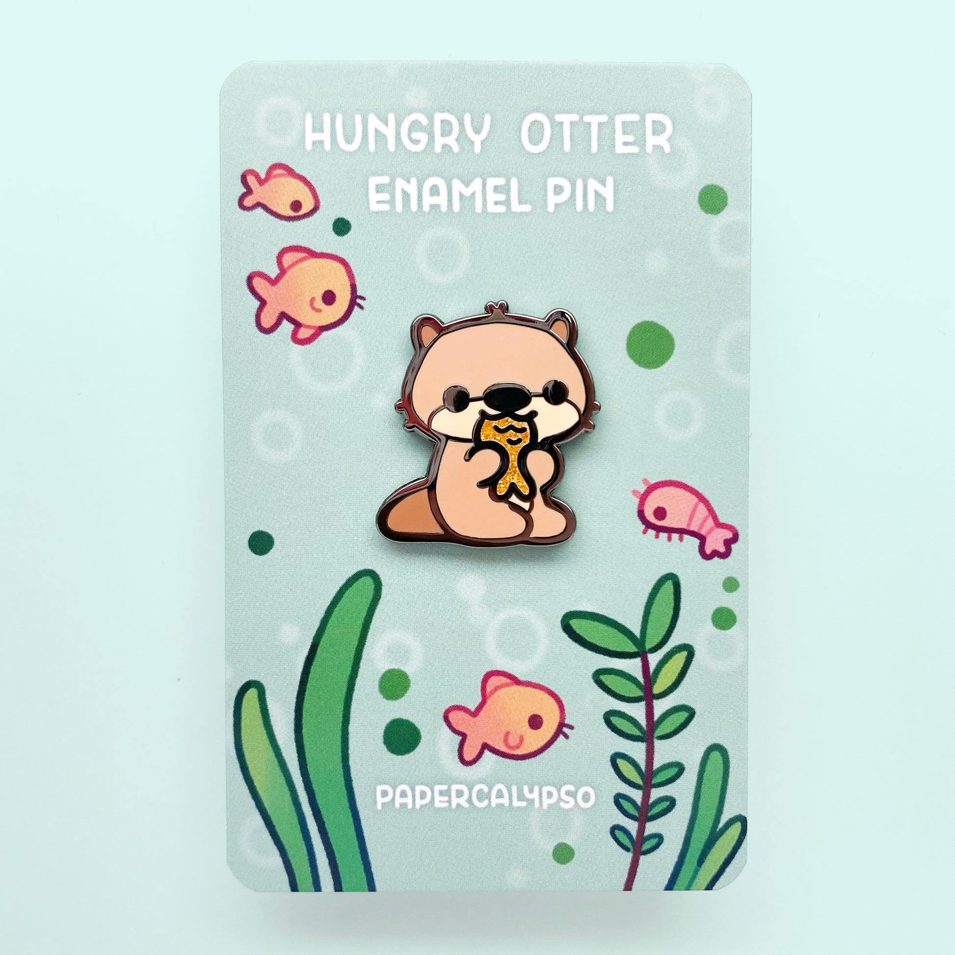 A black nickel hard enamel pin of a brown river otter baby eating a glittery orange fish. He's light brown and has cute whiskers.