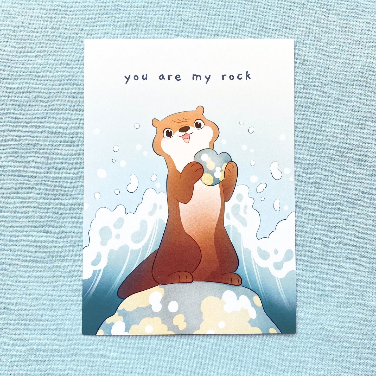 the card is a drawing of a european river otter holding a rock and smiling. It's standing on a boulder and the water crashes behind him, making a big wave splash in the background. On the top is the text "You are My Rock"