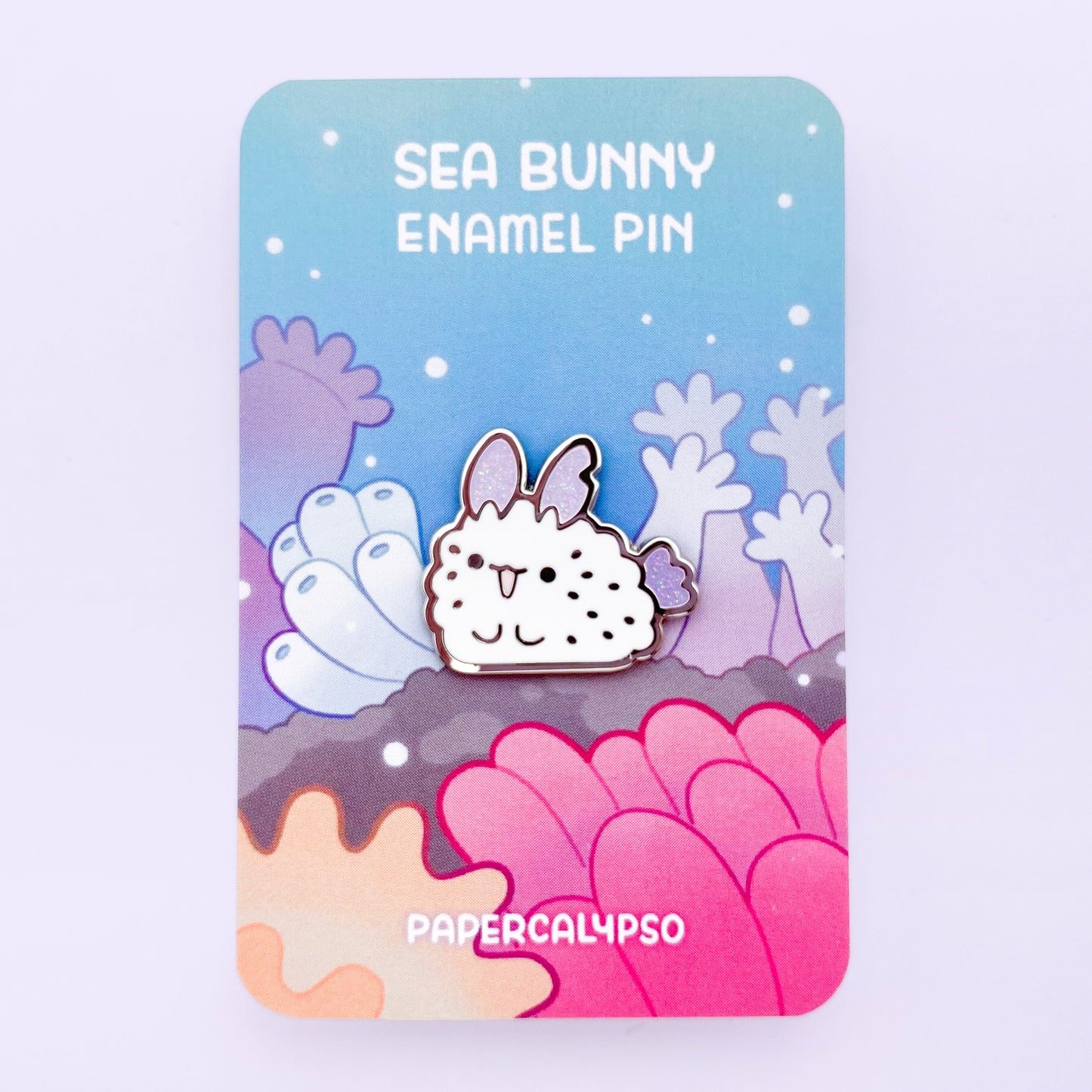 A nickel plated hard enamel pin of a sea bunny (jorunna parva) chilling in the coral reef. The gills and ears (rhinophores) are purple and glittery.