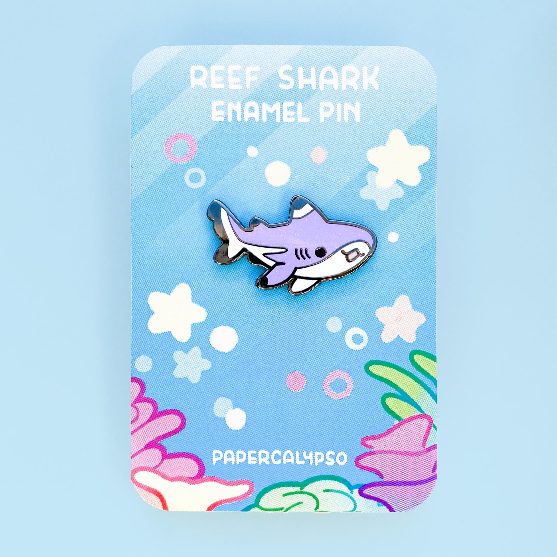 A black nickel hard enamel pin of a glittery purple blacktip reef shark. He has a white tummy and black enamel fin tips. He's smiling with his mouth open and has very cute proportions.