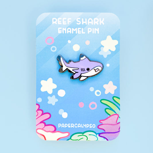 A black nickel hard enamel pin of a glittery purple blacktip reef shark. He has a white tummy and black enamel fin tips. He's smiling with his mouth open and has very cute proportions. 1